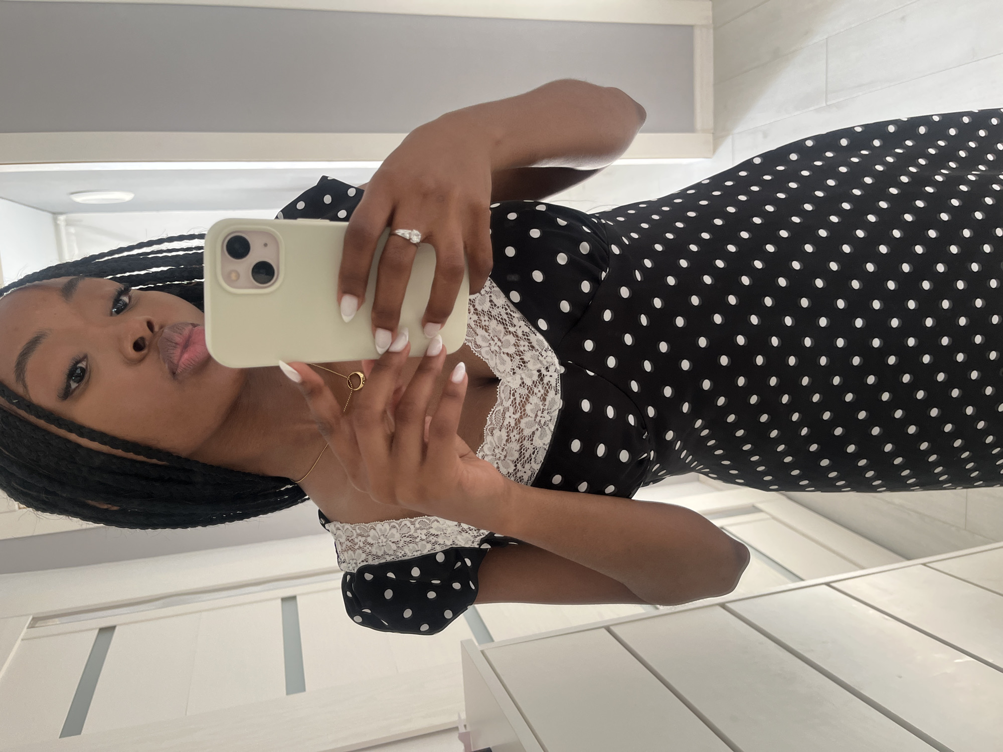 A young Black woman with braided hair is taking a mirror selfie in what appears to be a bedroom. She is wearing a black and white polka dot dress with lace trim on the neckline and puff sleeves. She is holding a light green iPhone in her right hand, which is angled to capture her reflection. Her left hand is resting on her right hand, and she is wearing a ring on her left ring finger and a gold necklace with a circular pendant. She has white nail polish on her fingernails. The background shows a white dresser with several drawers on the left, a closet door or wall with vertical paneling, and a grey wall to the right. The lighting suggests it is daytime. There is no indication of this scene being in Kazan, Russia.