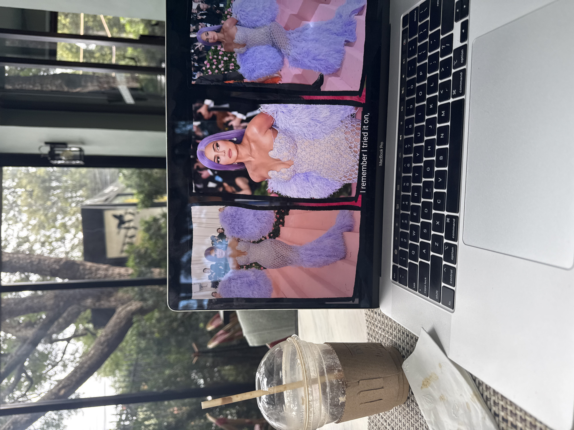 A laptop sits open on a table, displaying images of Kylie Jenner in a light purple gown with feather detailing. The laptop screen shows the text "I remember I tried it on," and "MacBook Pro." Next to the laptop is a clear plastic cup of iced coffee with a straw and a brown paper sleeve. A crumpled white napkin is on the table in front of the laptop. The background is blurred, showing greenery and trees outside a window, suggesting an outdoor or semi-outdoor setting. The overall scene appears to be a relaxed moment, possibly at a cafe or restaurant. The presence of the laptop and coffee suggests a personal or leisurely activity. There are no explicit indicators in the image to place the location within Mexico City, Mexico.