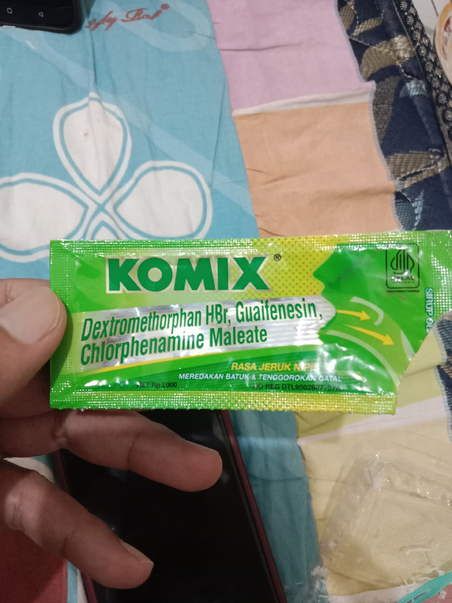 A person's hand holds a small, bright green sachet of Komix Dextromethorphan HBr, Guaifenesin, Chlorphenamine Maleate syrup. The sachet is partially opened, revealing a yellow checkered pattern along the bottom edge. The packaging is designed for a cherry flavor, indicated by "RASA JERUK." In smaller text, it states "Meredakan Batuk & Tenggorokan Gatal" (Relieves Cough & Sore Throat) and "No. Reg. DTL9502807737A." A "HALAL" certification logo is visible in the top right corner. The background consists of a patterned fabric, possibly a bedsheet, with a light blue and white floral design. The overall scene appears to be indoors, with hints of other materials and objects in the background suggesting a domestic environment, potentially in Brangsong, Indonesia.