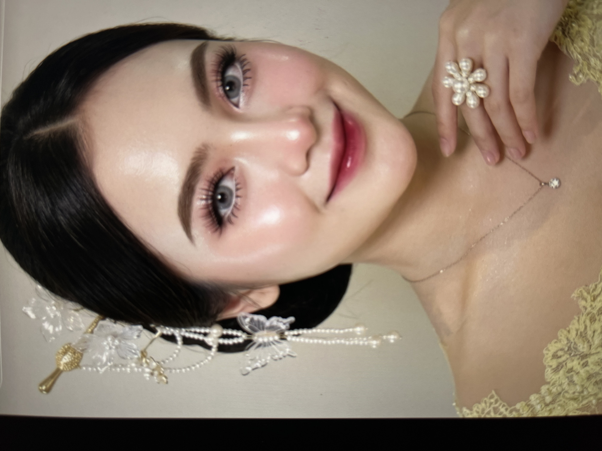 The image is a close-up portrait of a young East Asian woman with dark hair, styled with a golden and pearl hair accessory featuring butterfly embellishments. She is wearing elaborate makeup, including false eyelashes, pink eyeshadow, and dark pink lipstick. Her eyes are a light grey color, suggesting the use of contact lenses. She has on a necklace with a small diamond pendant and a large, floral-shaped ring adorned with pearls on her right hand. Her attire appears to be a yellow dress with lace detailing. The background is a plain, light-colored wall. The lighting is bright, highlighting the woman's features and the details of her accessories and makeup. There is no indication of the specific location within Cirahayu, Indonesia, or any environmental context beyond the studio-like setting.