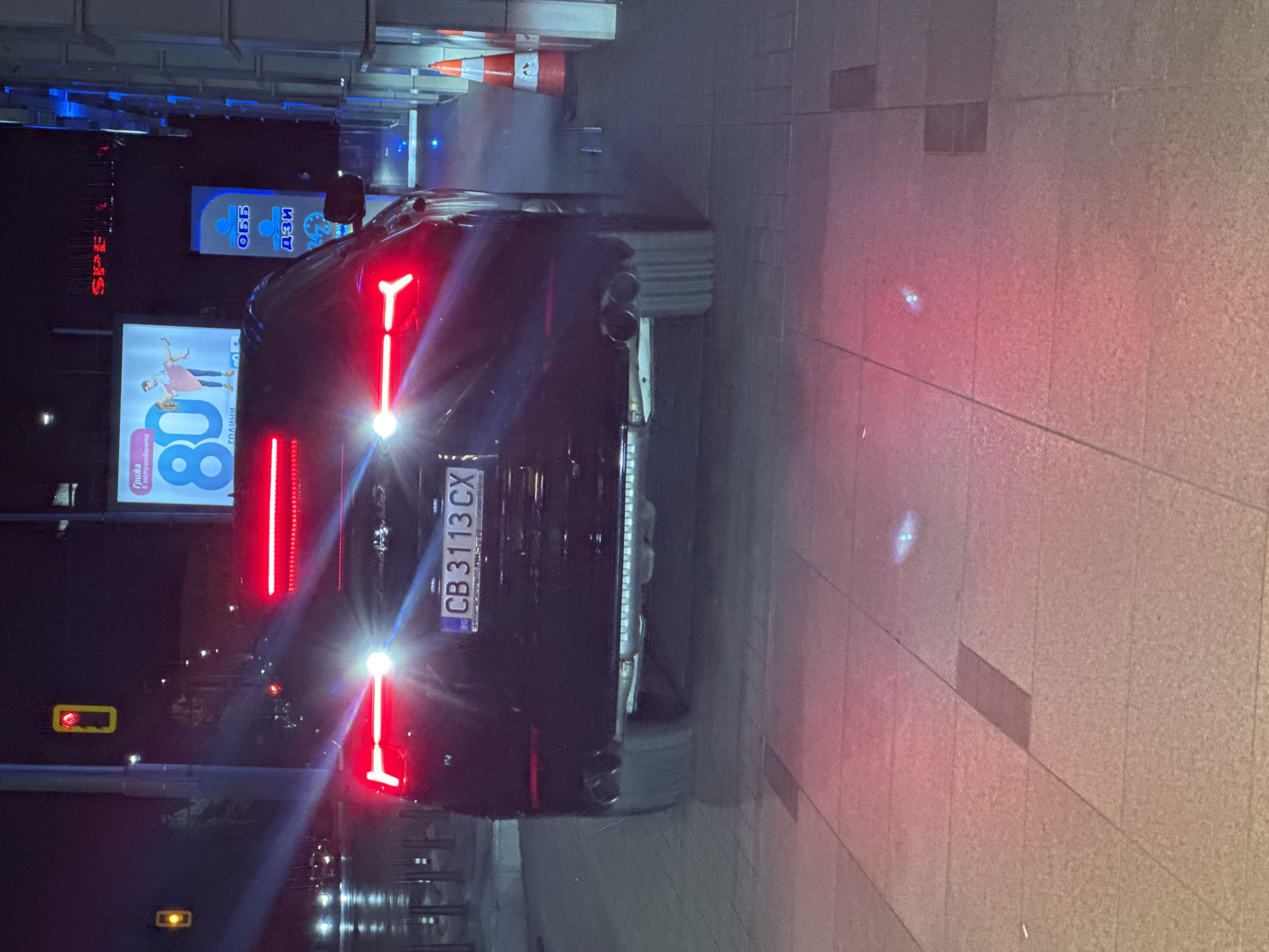 The image shows the rear of a black car parked on a sidewalk in what appears to be an urban setting. The license plate reads "CB 3113 CX," indicating registration in Sofia, Bulgaria. The car's taillights are illuminated, featuring a distinct vertical bar design. The vehicle is parked near a building with bright advertising billboards, including one that reads "80 ГОДИНИ" (80 years). Other signs and lighting fixtures are visible in the background. The ground is paved with rectangular tiles. There are no people visible in the immediate vicinity of the car. The scene is captured at night, with streetlights and illuminated signs contributing to the light levels.