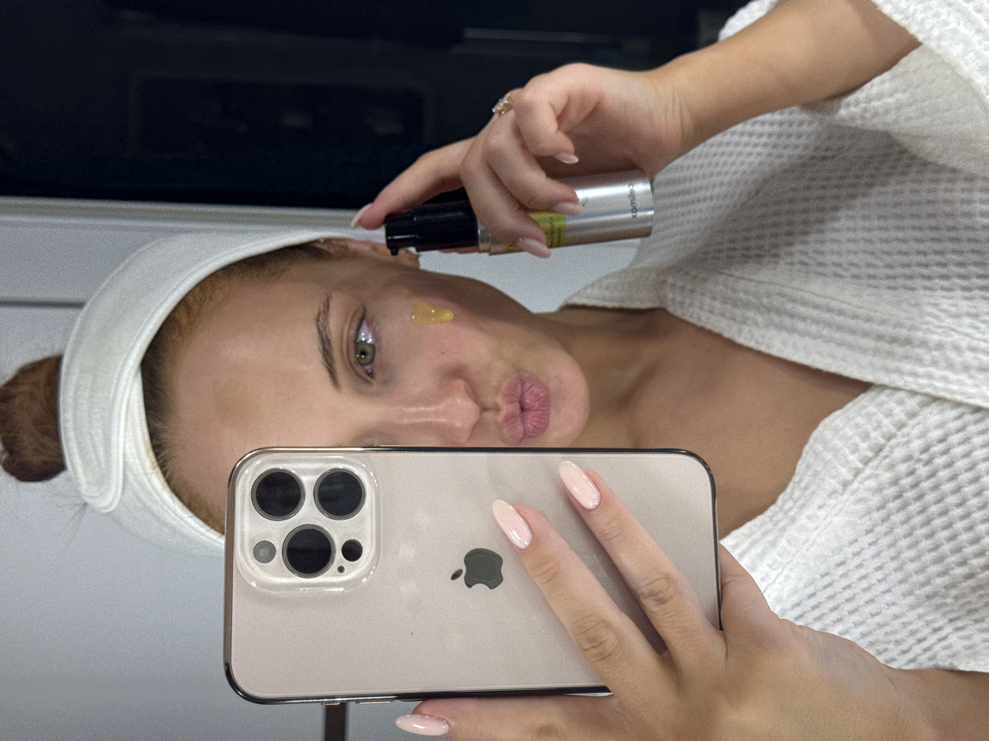 A woman wearing a white bathrobe and headscarf is applying a serum to her face, as she takes a selfie with an iPhone. A drop of the serum, a yellow liquid, is visible on her cheek. The serum's packaging is labeled "xorilos." The woman's expression is a playful pout. The location, Chaikovsky, Russia, cannot be determined from the image.
