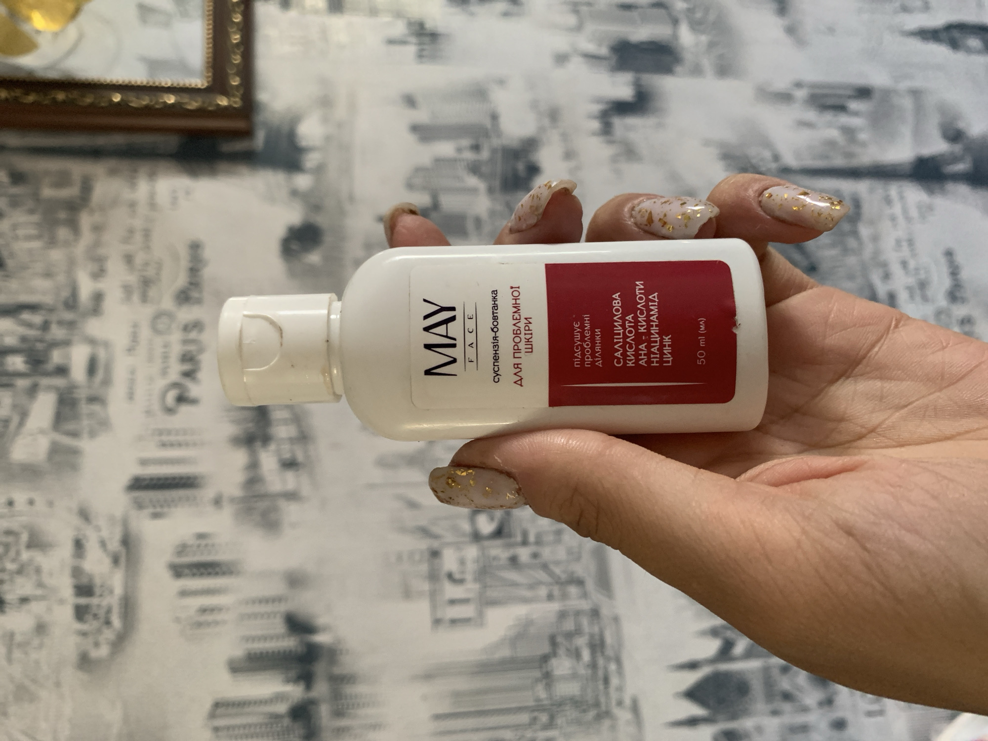 A person is holding a white bottle of MAY FACE suspension for problematic skin. The bottle has a white cap and a label with text in Ukrainian. The label is primarily white with a red section at the bottom. The text on the red section, from top to bottom, reads "підсушує проблемні ділянки" (dries problem areas), "САЛІЦИЛОВА КИСЛОТА" (salicylic acid), "АНА - КИСЛОТИ" (AHA - acids), "НІАЦИНАМІД" (niacinamide), "ЦИНК" (zinc), and "50 ml (мл)" (50 ml). The person's hand and nails, which are decorated with gold flakes, are visible in the foreground, partially obscuring the bottle. The background is a wallpaper with a repeated pattern of cityscapes, seemingly Paris, with buildings and script. The lighting is natural and appears to be coming from the right.