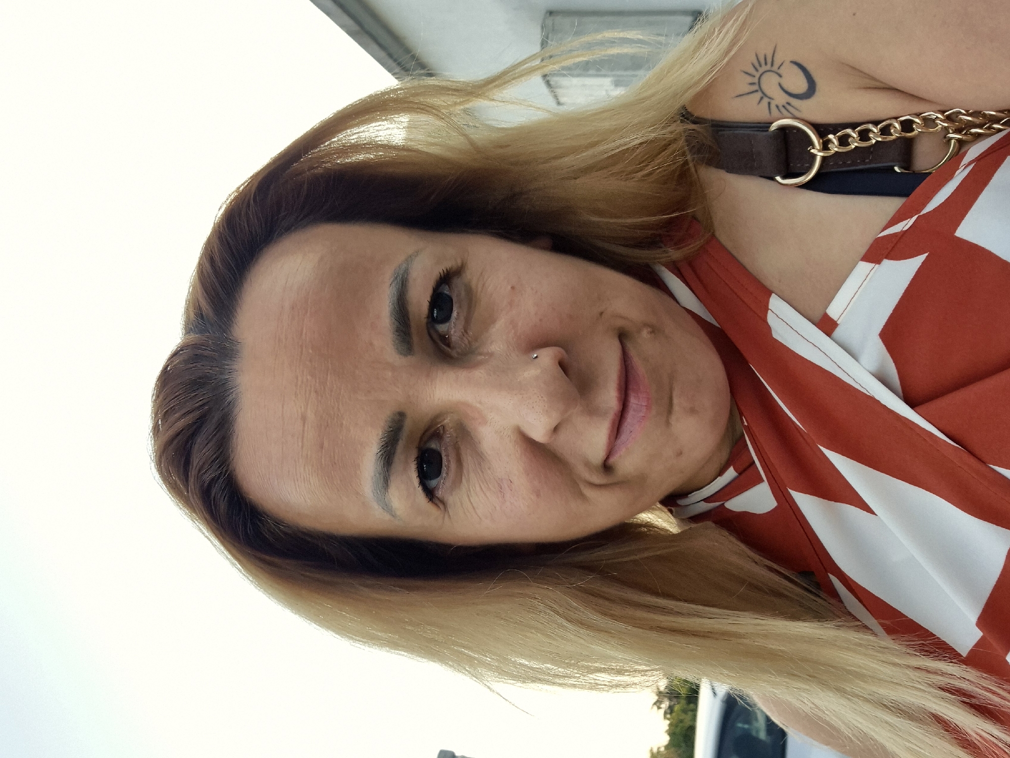 A close-up, head-and-shoulders selfie of a woman with blonde hair, light skin, and brown eyes is depicted. She is wearing a red and white patterned top. She has a small nose ring. A tattoo of a sun and crescent moon is visible on her right shoulder. A purse strap with a gold chain is also visible. The background is blurred, showing a white building and some greenery.