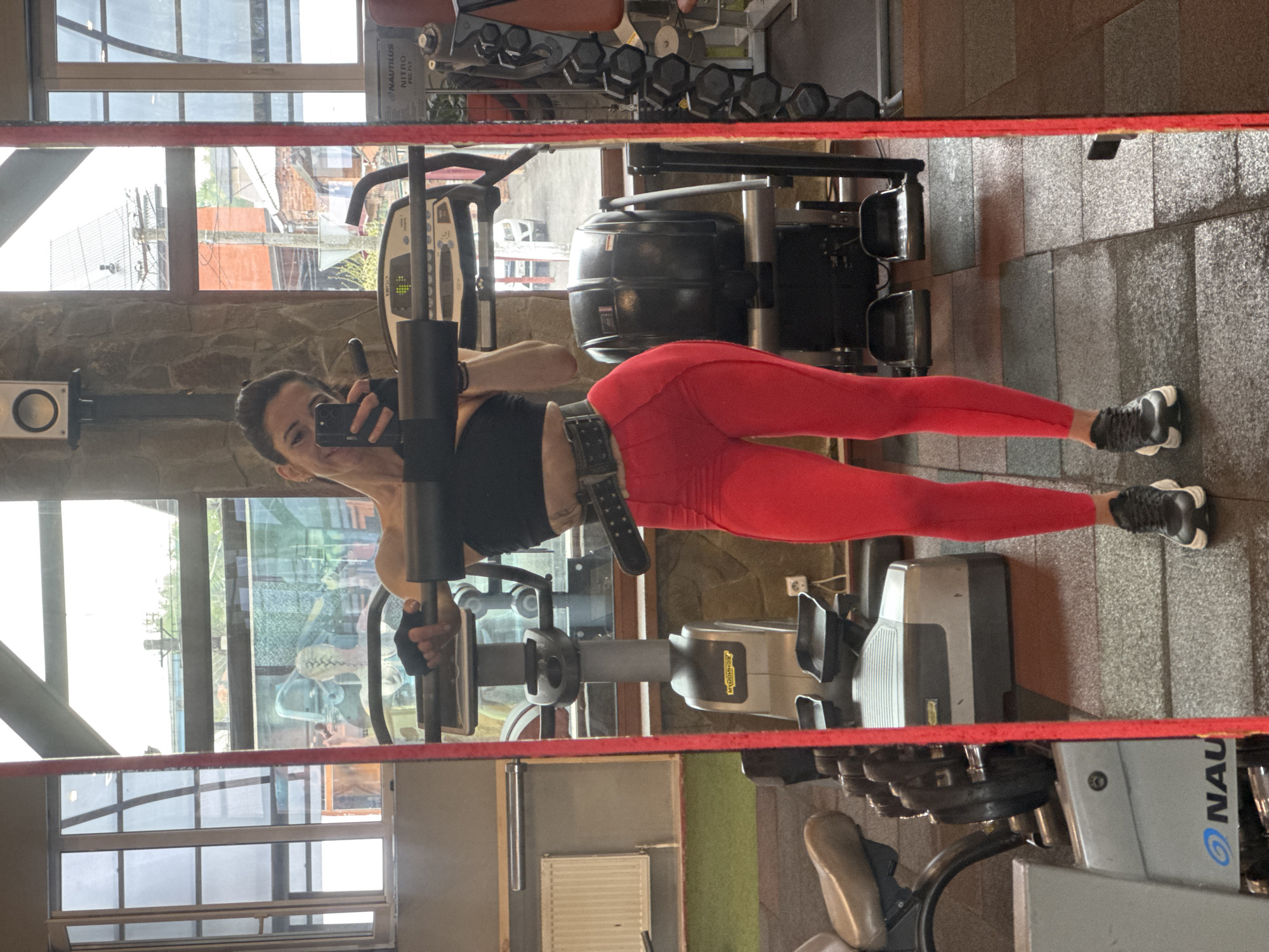 A woman is reflected in a mirror at a gym in Vladikavkaz, Russia. She wears black workout top, red leggings, grey sneakers, and a weightlifting belt. She is holding a phone, presumably taking a photo of herself. The gym includes Nautilus Nitro and Technogym equipment. Dumbbells are visible in the background. The gym has large windows that look out onto the city.