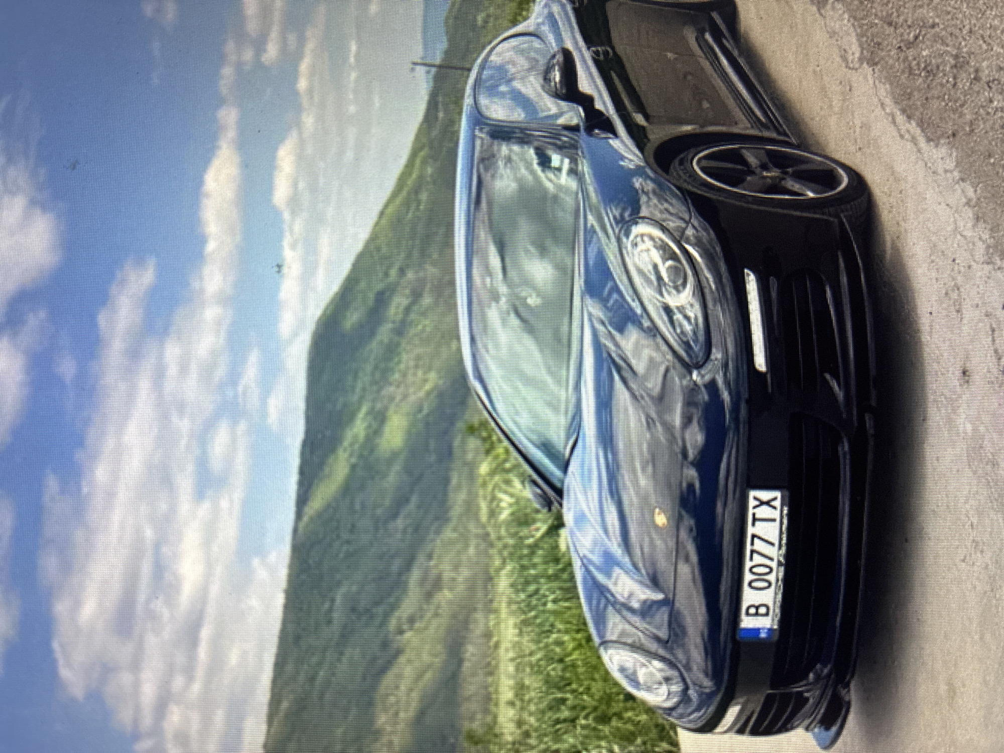 A dark-colored Porsche sports car is prominently featured, parked on a paved surface, likely a roadside or scenic overlook. The car is angled towards the viewer, with its sleek, reflective surface reflecting the bright sky and surrounding landscape. In the background, a vast, lush green mountain range stretches under a clear blue sky adorned with white, fluffy clouds, suggesting a beautiful day. The setting is consistent with mountainous areas around Asenovgrad, Bulgaria. The time of day is daytime, indicated by the bright, natural lighting. The car is stationary, with no specific activity taking place other than being parked. A notable detail is the front license plate, which clearly displays the registration "B 0077 TX". The "B" prefix on the license plate further supports the location being Bulgaria. No people are visible in the image. The overall composition highlights both the luxury vehicle and the picturesque natural scenery.