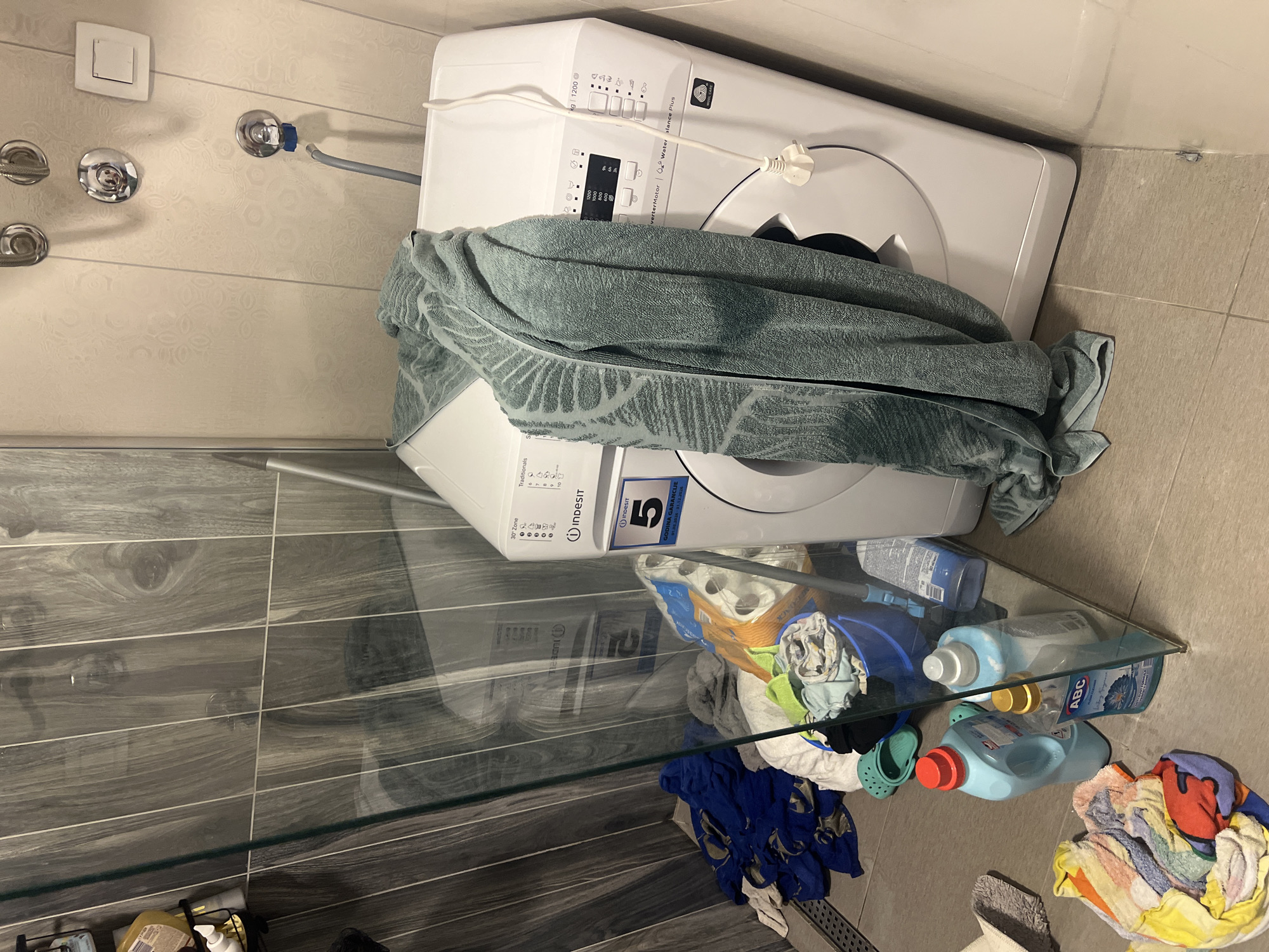 The image shows a top-down view of a bathroom or laundry area. A white washing machine is positioned in the upper part of the frame, partially covered by a large, folded green towel. Underneath the washing machine, a transparent glass shelf is mounted. The shelf holds various laundry-related items, including detergent bottles and other cleaning supplies. Below the shelf, a pile of assorted clothing and fabrics is scattered on the floor. The bathroom walls are tiled with a beige patterned tile. The floor is covered in grey wood-like tiles. The location, Budva, Montenegro, cannot be derived from the image content alone.