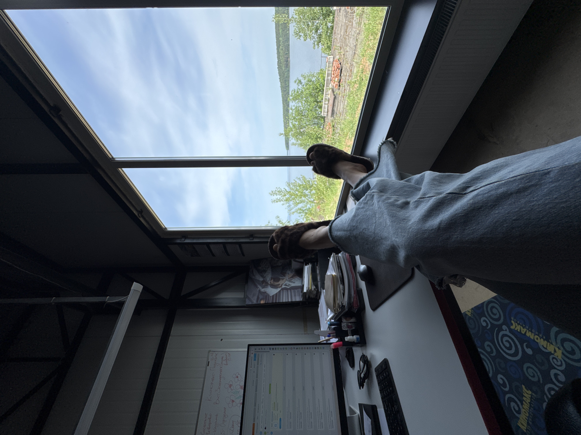 A person in a gray robe and brown slippers is relaxing with their feet up on the edge of a desk, near a window with a view of a lake and trees. The scene is taken from the perspective of the person looking out the window. The desk contains a computer monitor, keyboard, mouse, papers, and other office supplies. A whiteboard with writing on it is visible on the wall behind the desk. The window is positioned at an angle in the ceiling of what appears to be a prefabricated metal building. A mat with a spiral design reading "киномакс" is partly visible under the desk. The whiteboard contains text that mentions "Сосенское", "Говорово", "Киномакс Подолька", "Казань", and "Вист Зеленодольском". I do not have information to determine the location within the city of Chaikovsky, Russia.