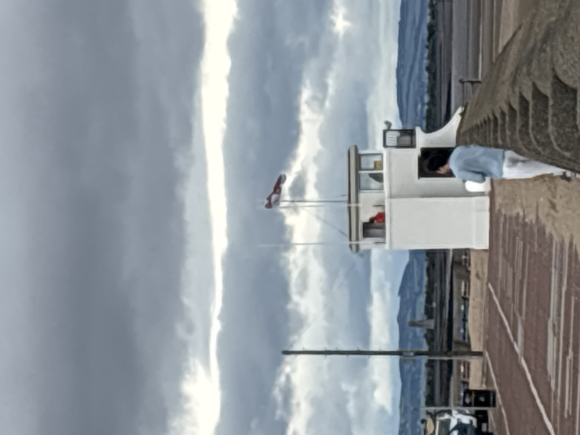 The image depicts a vertical scene featuring a white lifeguard tower against a backdrop of dramatic, cloudy skies. The tower has a small enclosed upper level with a window, through which a person wearing a light blue top and white trousers is visible, leaning forward as if looking down. A flag with red and white markings is attached to a pole in front of the tower. The foreground shows a textured, reddish-brown surface, possibly a pier or boardwalk, with a dark railing on the right side. In the distance, hills or mountains are faintly visible under the overcast sky, suggesting a coastal or lakeside setting. The overall impression is of a possibly breezy or stormy day, given the heavy cloud cover. The scene appears to be set during the daytime, with diffused light. There are no visible text elements in the image.