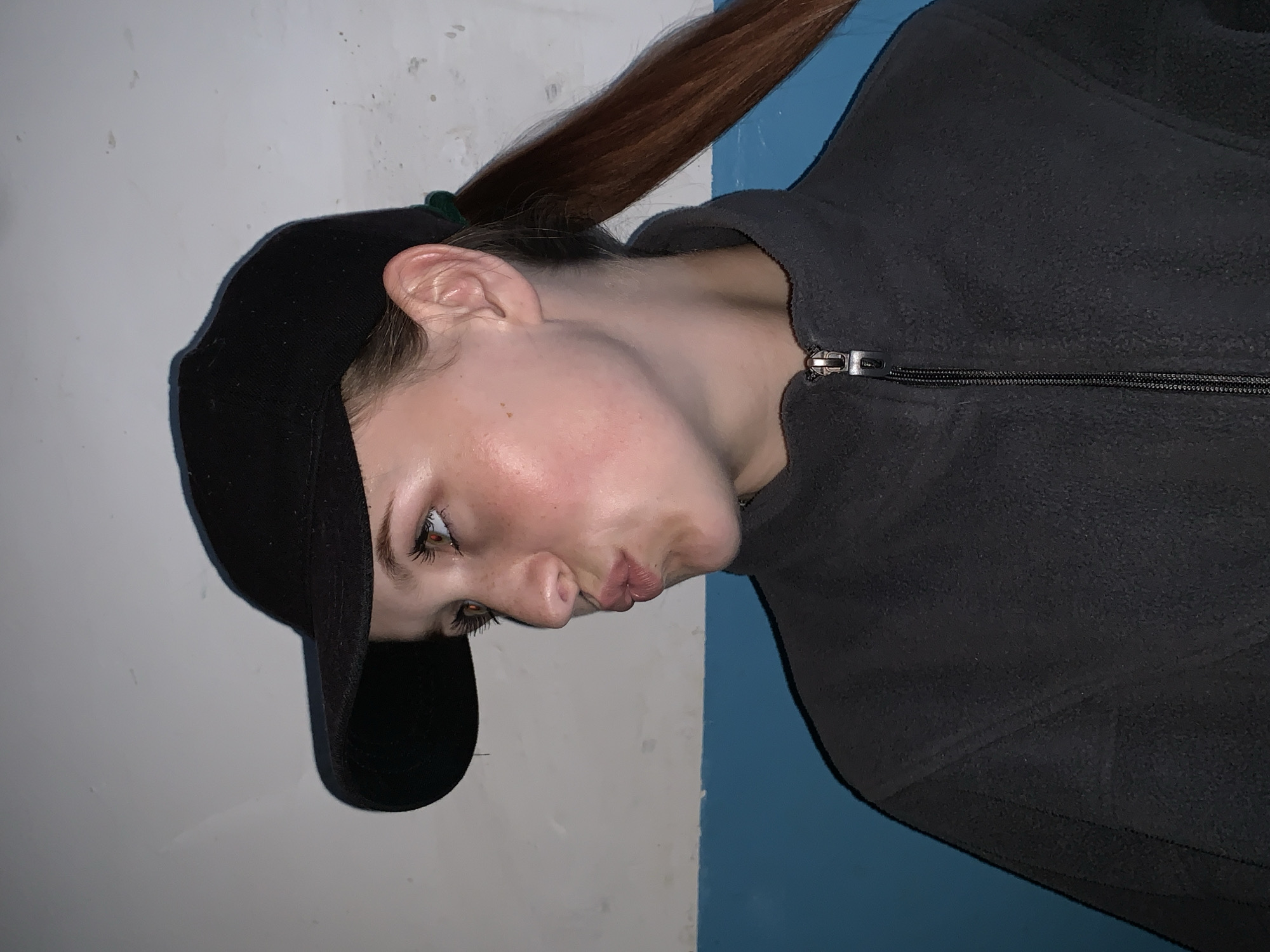 A woman wearing a black baseball cap and a gray fleece jacket stands in front of a wall that is painted white and blue. Her brown hair is pulled back in a ponytail. Her expression is neutral. The location is not discernible from the image.