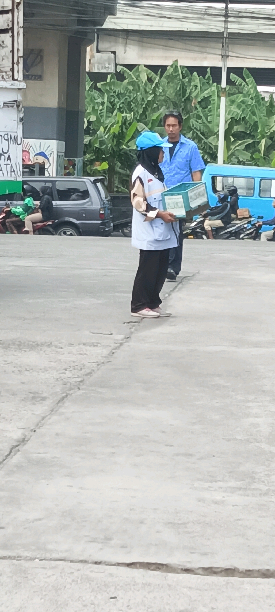 The image captures a street scene in East Jakarta, Indonesia, on a day with overcast skies. In the foreground, a person wearing a blue cap, hijab, and a light blue vest over a patterned shirt stands holding a light blue box. They are wearing black pants and light pink shoes. To their right, a man in a blue shirt stands facing slightly away from the viewer.
In the background, various elements suggest a public area. To the left, part of a building with a mural is visible, alongside a parked gray minivan. Further back, several motorcycles are parked, with some people on or near them. A bright blue vehicle, possibly a bus or van, is also present. Lush green banana trees form a dense backdrop to the scene. The ground is paved with concrete, showing visible cracks and seams.
