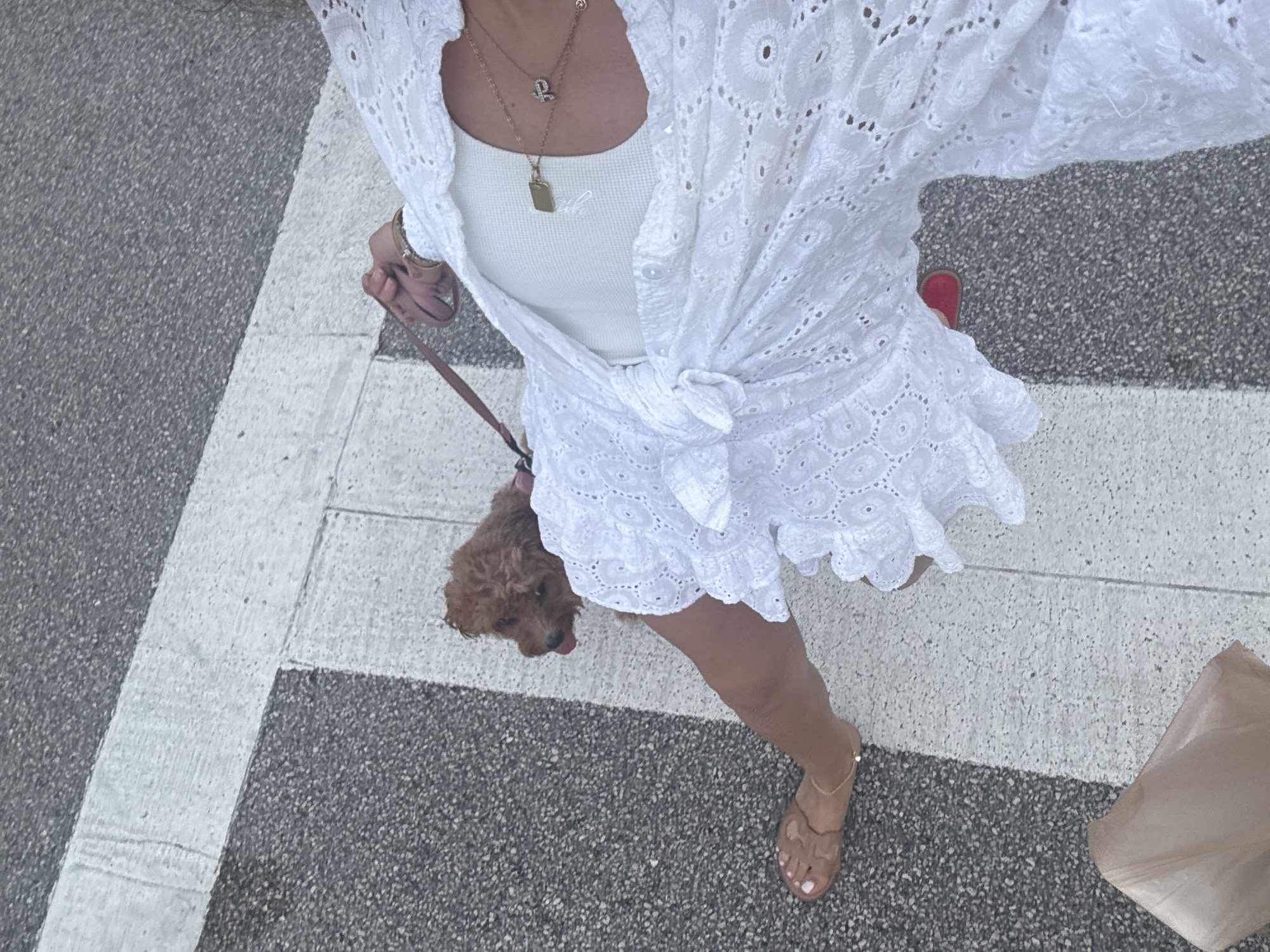 A high-angle photograph shows a woman and a dog standing on a crosswalk in Boca Raton, Florida, USA. The woman is wearing a white eyelet shirt tied at the waist over a white tank top, with a matching white eyelet skirt, and brown sandals. She is wearing two necklaces. A brown poodle is on a pink leash held in her left hand. A brown shopping bag is on the ground to the lower right of her. The crosswalk is made of asphalt between parallel white stripes.