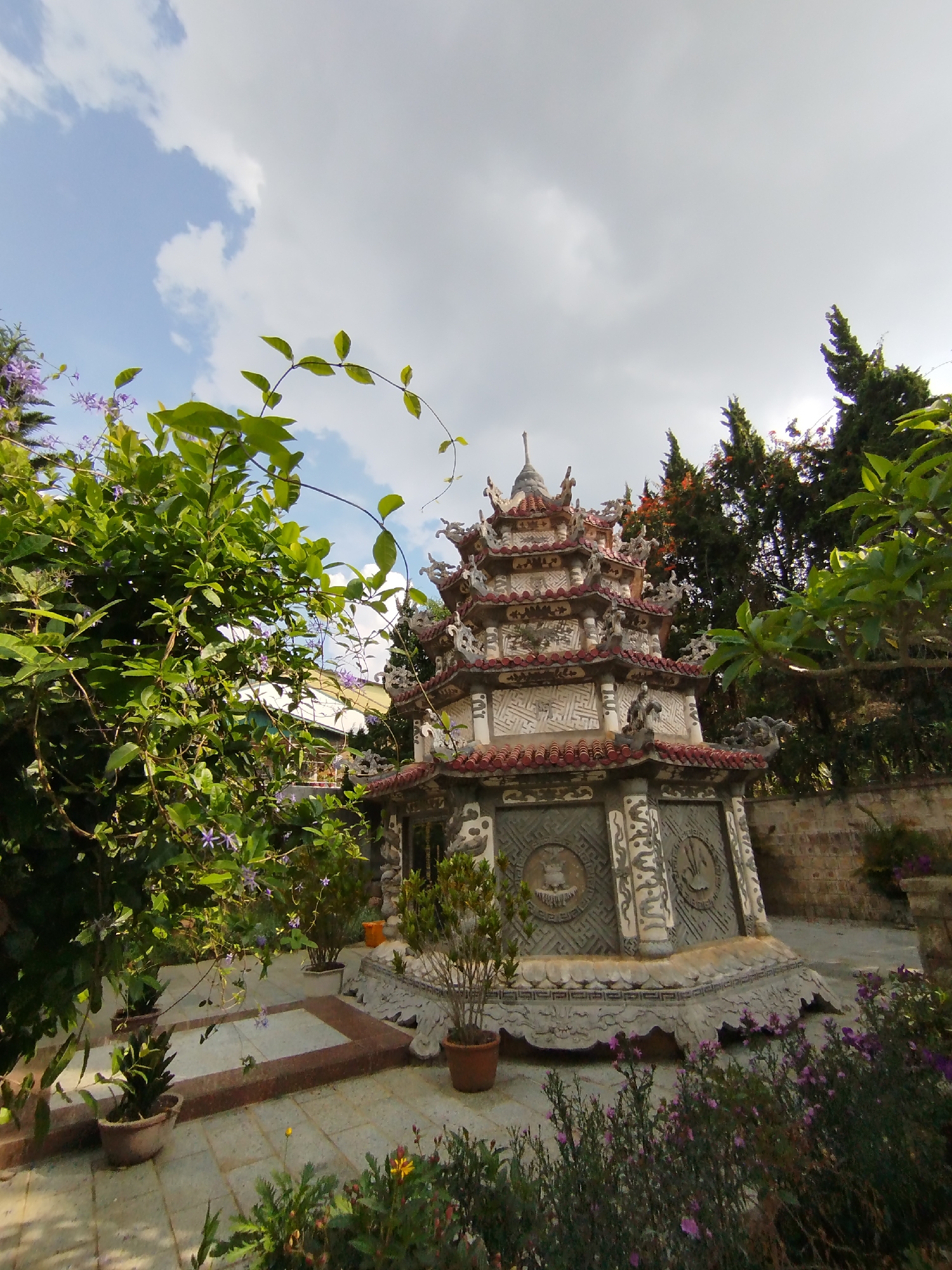 An outdoor, low-angle photograph captures an ornate, multi-tiered stone structure resembling a small pagoda or stupa, set within a lush garden under a partly cloudy sky. The structure, positioned slightly to the right of the frame's center, is intricately carved with detailed relief work, featuring multiple red-tiled roofs and stylized decorations on its eaves and facade. Its base appears octagonal or multi-faceted, with prominent vertical columns and circular motifs embedded in its sides.
The foreground on the left is dominated by vibrant green foliage, including a large bush and trailing vines adorned with small purple flowers, possibly framing the scene. Various potted plants are arranged around the base of the stone structure, with more green shrubs and purple-flowered plants visible in the bottom right. In the background on the right, tall, dark evergreen trees rise towards the sky, and a stone or brick wall is partially visible behind the main edifice. The ground is paved with light-colored tiles, forming a patio or walkway.
The sky transitions from bright blue with white clouds on the left to a more overcast, grey appearance on the right. The natural, soft lighting indicates daytime, likely morning or late afternoon. The scene is devoid of people or discernible activity, presenting a peaceful and decorative garden setting with cultural architectural elements. The location is Cam Ly - Đà Lạt, Vietnam. No visible text is present in the image.
