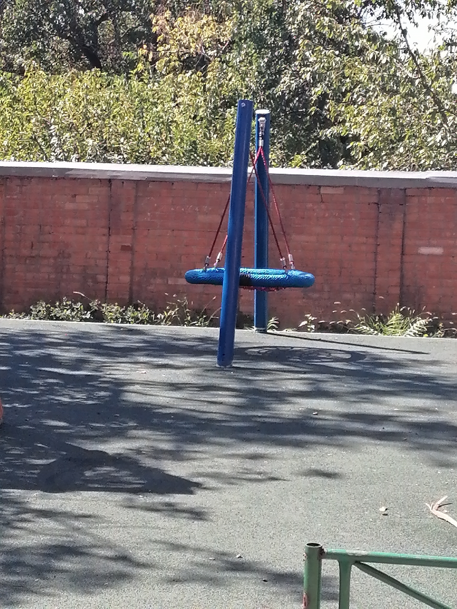 The image depicts a blue disc swing hanging from a metal A-frame structure in an outdoor playground. The swing is suspended by red ropes. Behind the playground equipment is a weathered brick wall, and beyond that, lush green trees. The ground of the playground is a dark, textured surface, likely rubberized safety surfacing, with tree shadows cast upon it. A green metal fence is partially visible in the foreground. The scene appears to be set on a sunny day. The location is identified as Rostov-on-Don, Russia.