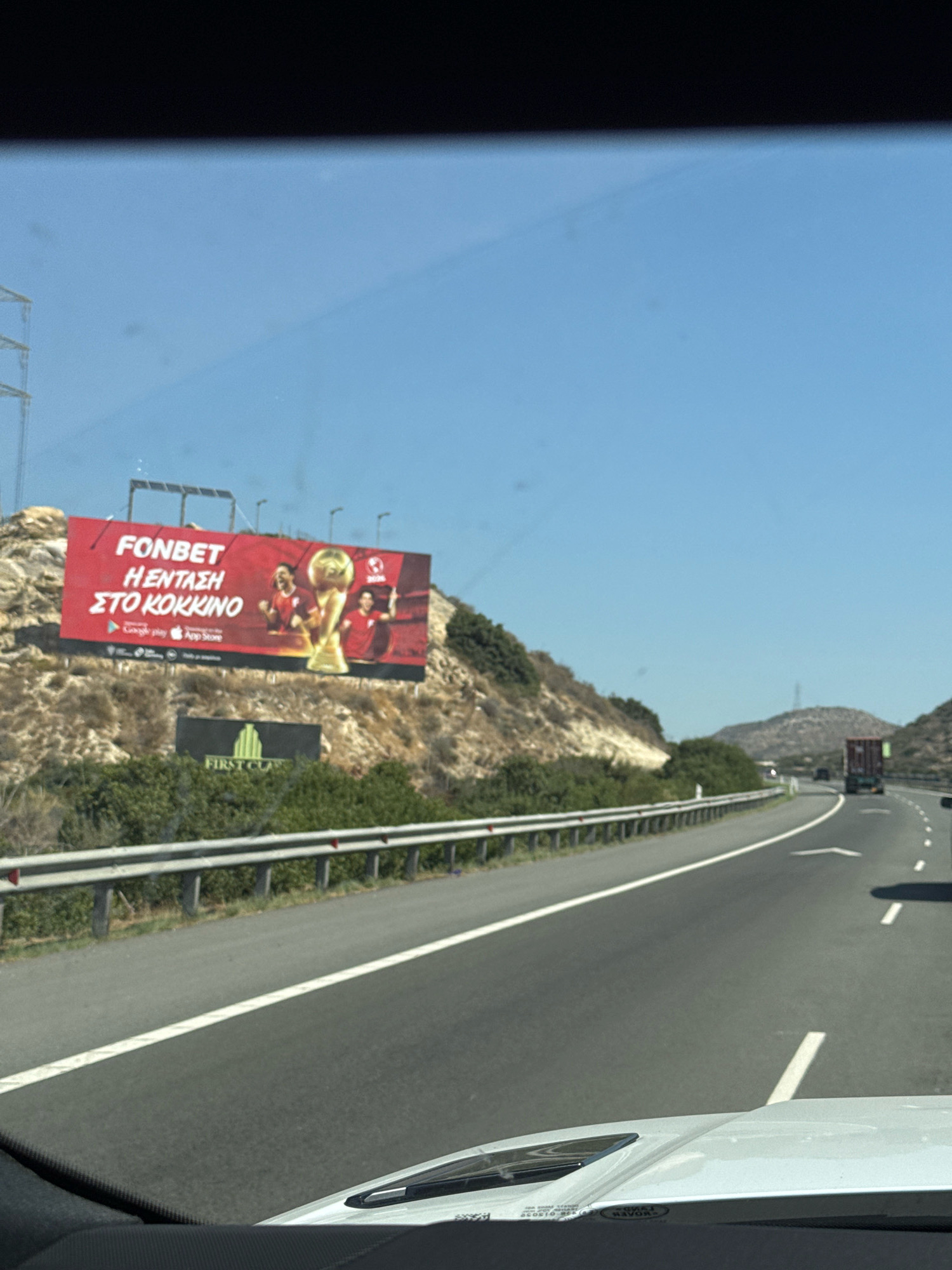 The image shows a billboard for Fonbet, a betting company, on the side of a highway in Psematismenos, Cyprus. The billboard features images of two men in red shirts holding a trophy, suggesting a sports betting advertisement. Below the main advertisement, a smaller billboard for "First Class" is visible. The highway is a multi-lane road with a metal guardrail along the side. In the distance, a truck and other vehicles can be seen on the road, and the landscape is characterized by dry, rocky hills with sparse vegetation. The sky is clear and blue, indicating a sunny day. The photograph appears to be taken from inside a moving vehicle, as evidenced by the dashboard and windshield in the foreground.