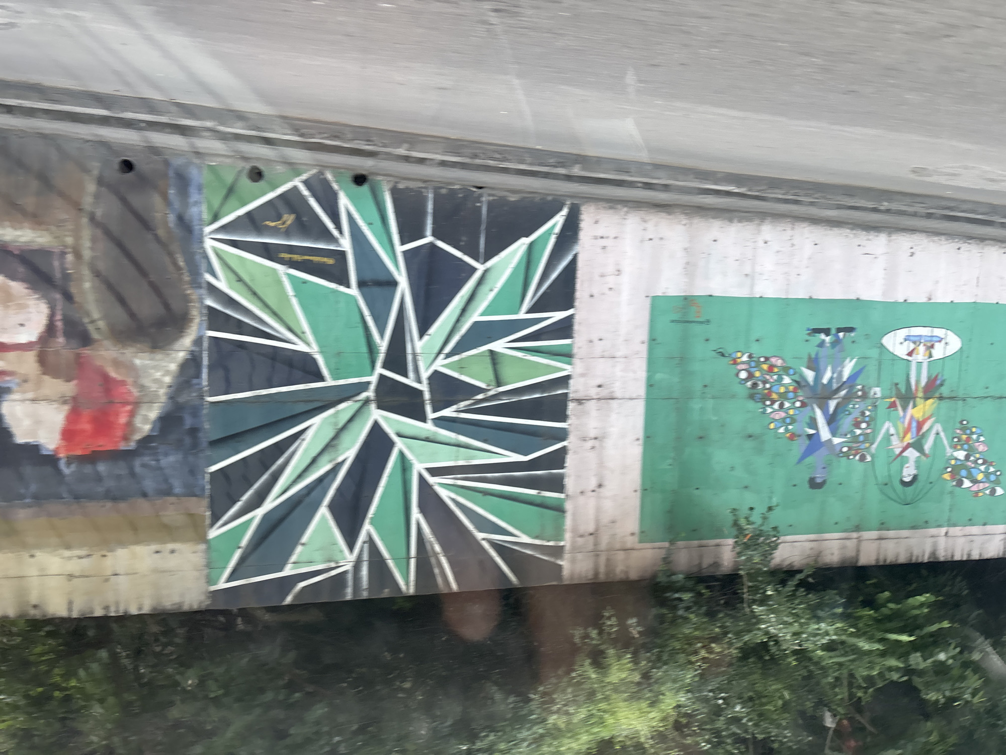This is a photograph taken from a moving vehicle on a street in Tbilisi, Georgia. The image features a concrete wall adorned with three distinct murals. The leftmost mural is on a green background and depicts two stylized figures holding umbrellas over their heads, with arrays of colorful eyes scattered around them. The middle mural is a geometric abstract design in shades of green, black, and white. The rightmost mural is partially visible and appears to be a portrait or figurative work in earthy tones with red accents. The wall is situated below a densely vegetated hillside, and a road with a concrete curb runs in front of it.