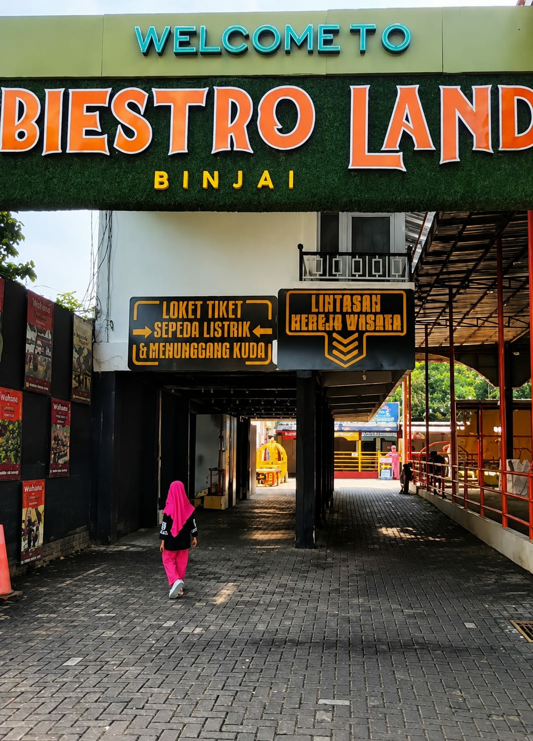 The image shows the entrance to "Biestro Land" in Binjai, Indonesia. A large green sign above the entrance reads "WELCOME TO" in teal lettering, followed by "BIESTRO LAND" in large orange block letters. Below this, the word "BINJAI" is written in smaller yellow letters.
To the left of the entrance, a dark-colored walkway leads further into the park. A young girl wearing a bright pink hijab and pink pants is walking away from the camera down this walkway. On the wall to her left, several posters advertising attractions are visible.
Above the walkway, two black signs with yellow text are mounted. The left sign, with an arrow pointing left, reads "LOKET TIKET SEPEDA LISTRIK & MENUNGGGANG KUDA" (Ticket Counter Electric Bicycle & Horse Riding). The right sign, with chevron-like arrows pointing down, reads "LINTASAN KERETA WISATA" (Tourist Train Track).
The right side of the image shows a covered walkway with a red railing, and what appears to be part of a tourist train track. Several people are visible in the distance, standing near the train track. The ground is paved with dark grey interlocking bricks. The environment is outdoors with sunlight casting shadows on the ground.