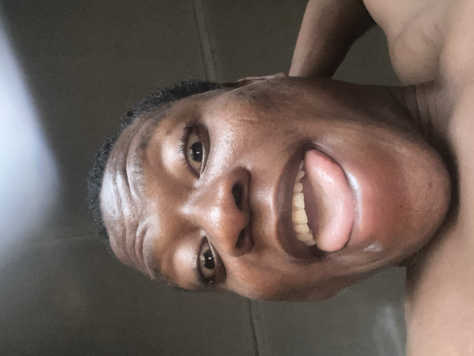 A close-up, slightly upward-angled shot depicts a dark-skinned individual with their tongue extended, appearing to make a playful face. The subject's eyes are open and directed towards the camera. The background is a blurred, neutral tone, devoid of specific details or landmarks. The city and location are unknown.