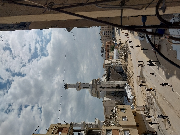 This image captures a street scene in Khan Yunis, Palestinian Territory, under an overcast sky with scattered clouds. The dominant feature is a partially damaged mosque located in the mid-ground, characterized by a large dome and a tall, white minaret with an ornate crown. Significant structural damage is evident on buildings flanking the street; a multi-story building to the left has extensively collapsed sections revealing rebar and interior structures, while a building fragment in the extreme foreground on the right also shows damage and exposed materials, acting as a partial frame.
The street itself is wide, unpaved or heavily covered in earth and rubble, with no visible lane markings. Piles of debris and dirt flank both sides of the thoroughfare. Approximately 25-30 individuals, including adults and several children, are scattered across the street and its immediate vicinity, walking in various directions. Most are solitary or in small groups of two or three. One person on the right side of the street appears to be pushing a small, wheeled cart. Overhead, a thin dark line, possibly a wire or cable, spans horizontally across the upper mid-section of the image. In the background, beyond the mosque, other multi-story residential or commercial buildings are visible, some appearing structurally intact while others show signs of possible damage or incomplete construction.