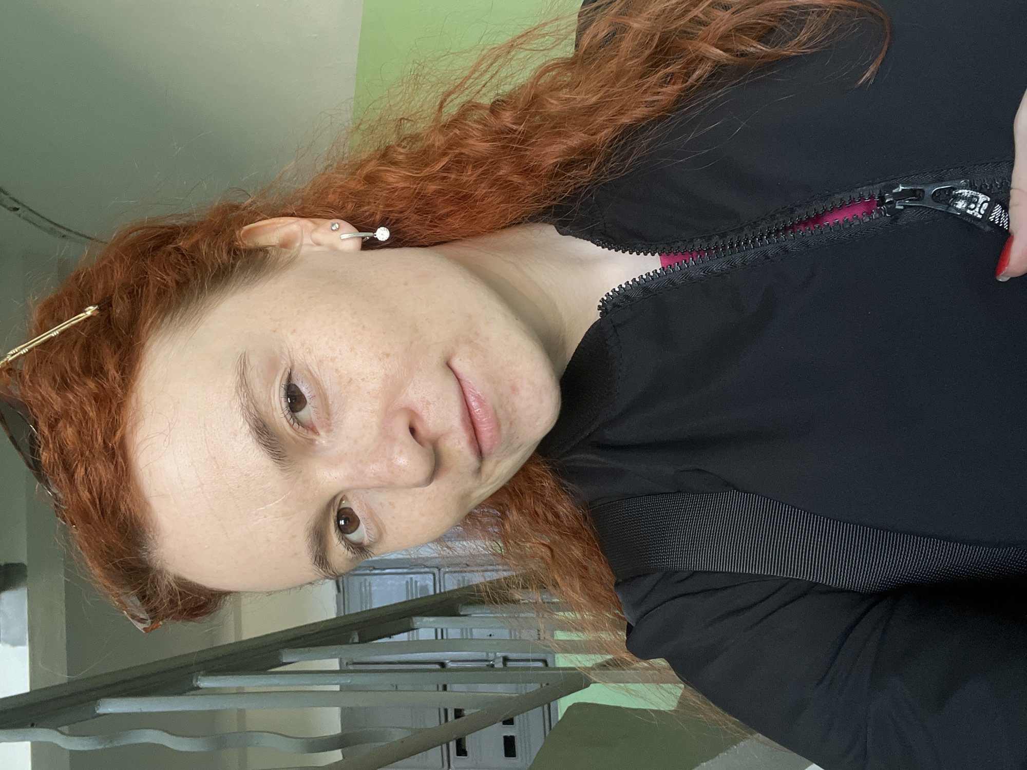 A woman with red, curly hair is standing on a staircase. She is wearing a black jacket, partially zipped up to reveal a pink lining, with a black strap over her shoulder. She has a silver earring in her left ear and a hair clip in her hair near her left temple. The background includes a light green wall and a gray staircase with metal railings. The picture was likely taken indoors in a building in Chaikovsky, Russia.