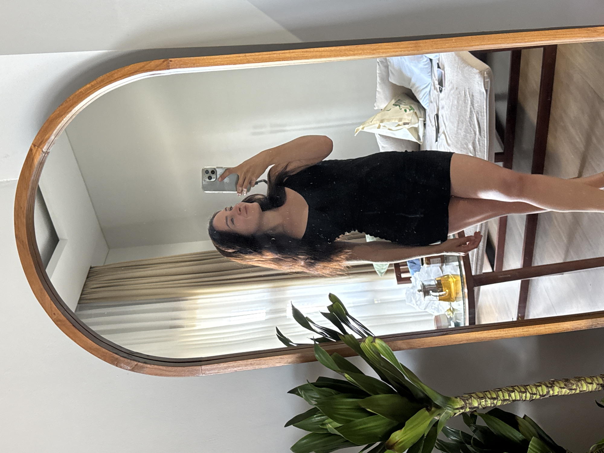 In North Kuta, Indonesia, a young woman with long, dark hair is taking a mirror selfie in a dimly lit room. She is wearing a black mini dress and is holding up her phone to capture her reflection. The mirror is framed by a wooden arch, and behind her, a couch with pillows and a table with a teapot can be seen. A large green plant stands to her left, and the room appears to be residential.