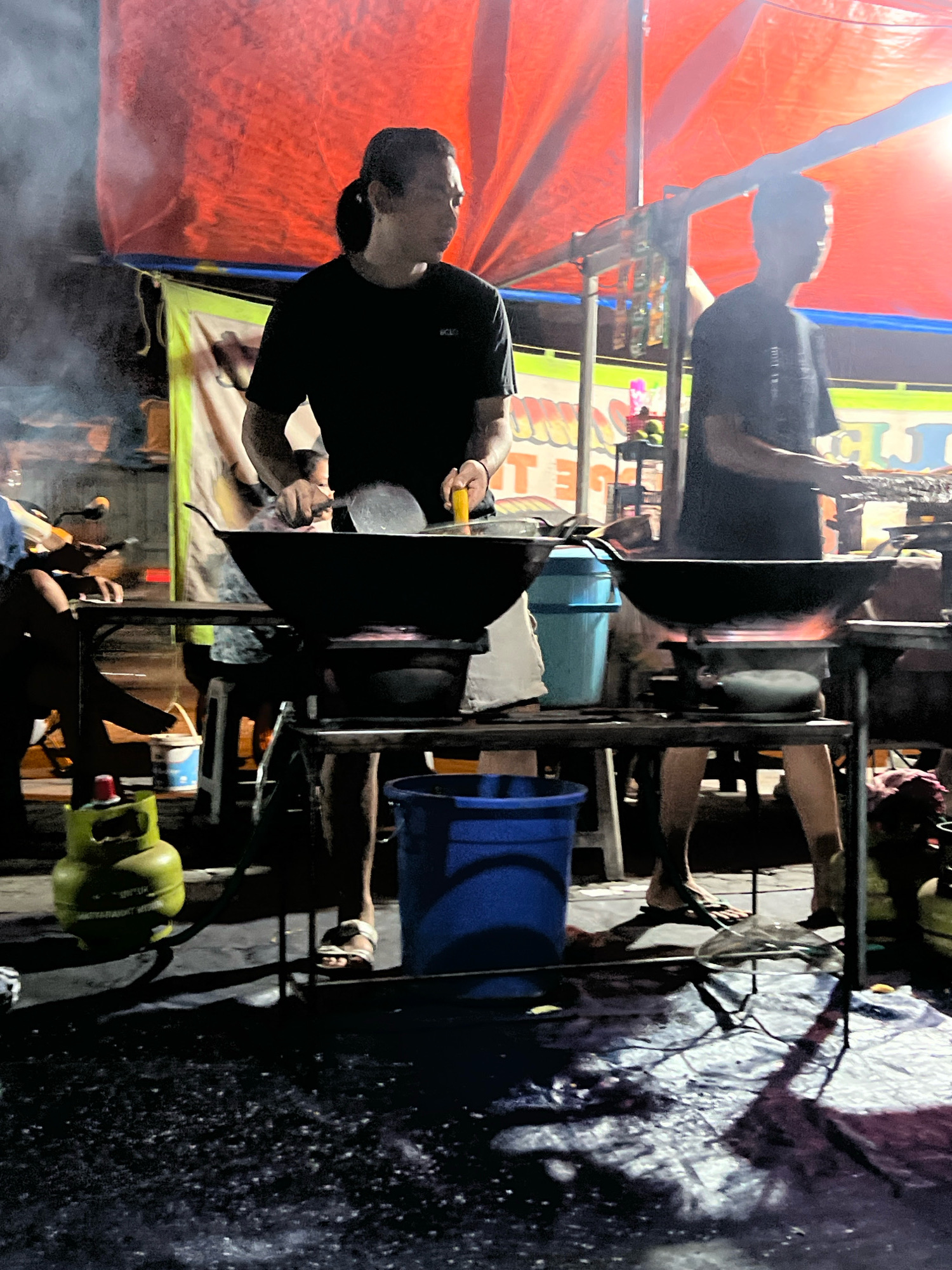 The image shows two men cooking outdoors at night in Tlogoadi, Indonesia. The men are standing behind portable cooking stations, each with a large wok over a stove. One man is wearing a black t-shirt and has his hair pulled back in a ponytail. He is using a spatula to stir food in the wok. The other man is wearing a black t-shirt and is also cooking. The background shows a red tent or canopy, a motorcycle, and what appears to be a street or marketplace setting. There are also propane tanks and buckets visible. The ground is wet and reflective, possibly from recent rain or water used for cleaning. The lighting is dim and atmospheric, with the cooking flames providing a warm glow.