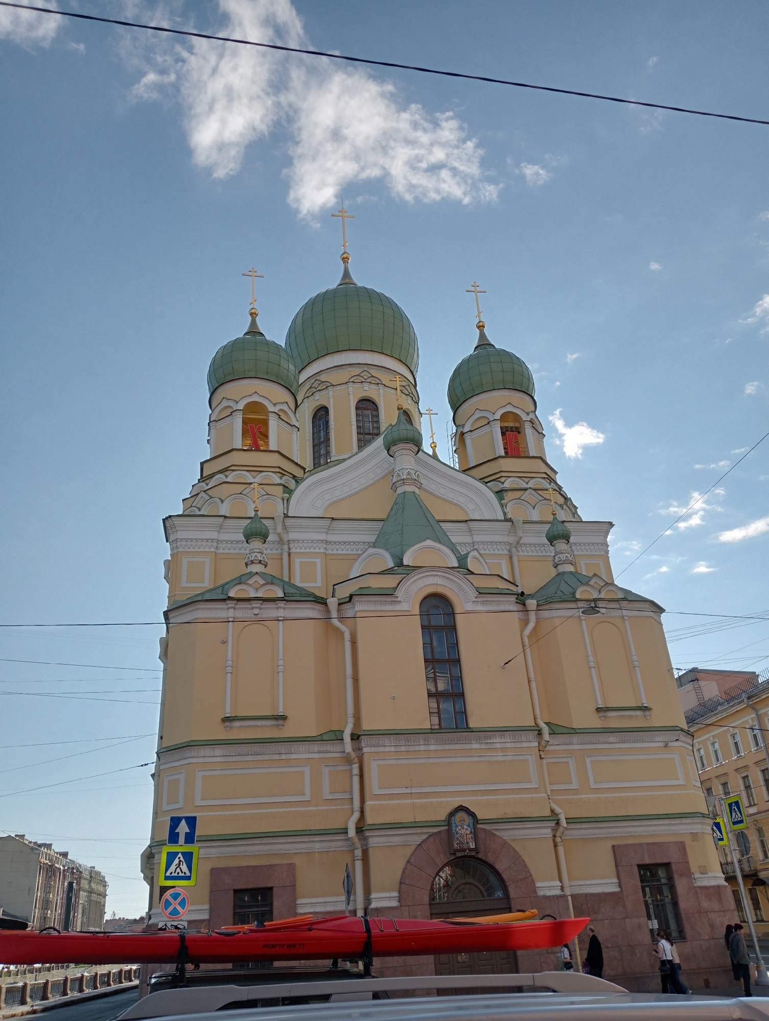 The image depicts the Church of the Assumption of the Holy Virgin in St. Petersburg, Russia, on a clear day with scattered clouds. The building is a prominent yellow structure with multiple green domes topped with golden crosses. The foreground shows the roof of a car carrying a red kayak. Several street signs are visible, including a pedestrian crossing sign and a directional arrow. In the background, other buildings and people can be seen. The church exhibits classical architectural elements with ornate detailing and arched windows. The sky is a vibrant blue, and power lines are visible across the top of the frame.