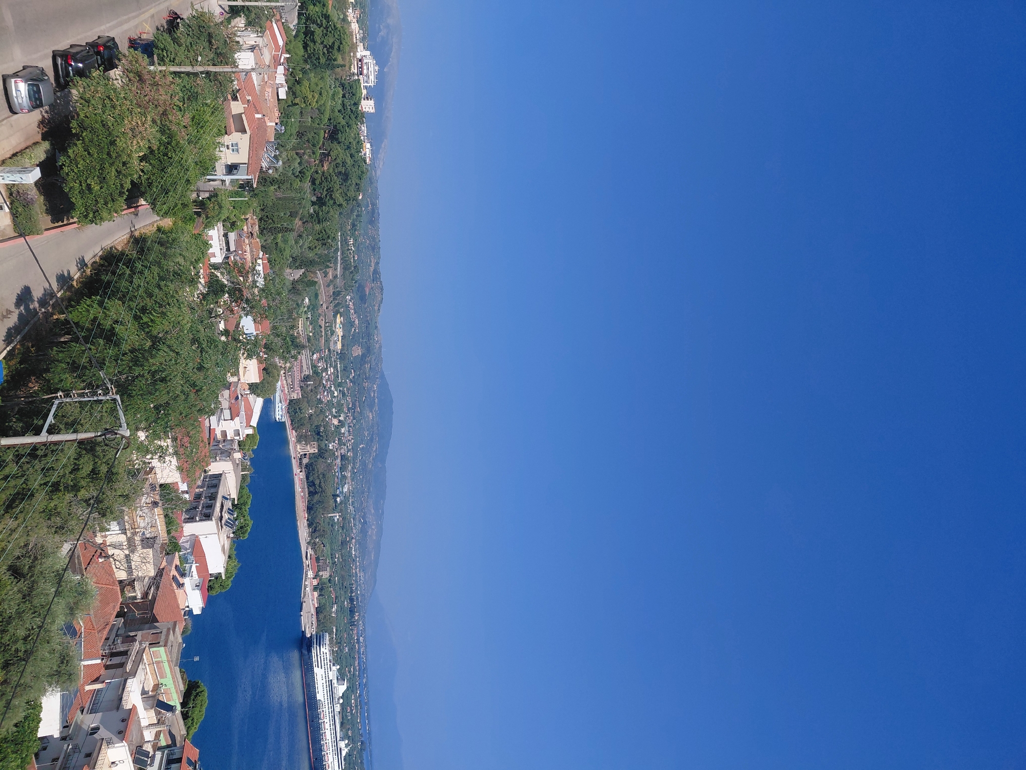 This is a high-angle, full shot of a coastal city on a clear, sunny day, likely in Municipal Unit of Aegio, Greece, given the characteristic red-tiled roofs and proximity to the sea. The dominant feature in the upper half of the frame is a vast expanse of clear, bright blue sky. Below the sky, a body of deep blue water, presumably the sea, occupies the right side of the midground. A large, white cruise ship with multiple decks and dark hull is docked in the water, close to the shore. The coastline is lined with buildings, many with red-tiled roofs, and appears to be a developed urban area. Behind the coastal buildings, a hilly landscape covered in dense green trees and scattered buildings extends into the background. The foreground depicts a street with parked cars and more residential buildings with red-tiled roofs, surrounded by lush green trees and vegetation. Power lines are visible crisscrossing through the scene. The overall impression is a sunny, Mediterranean coastal town. There are no people clearly visible in the image.