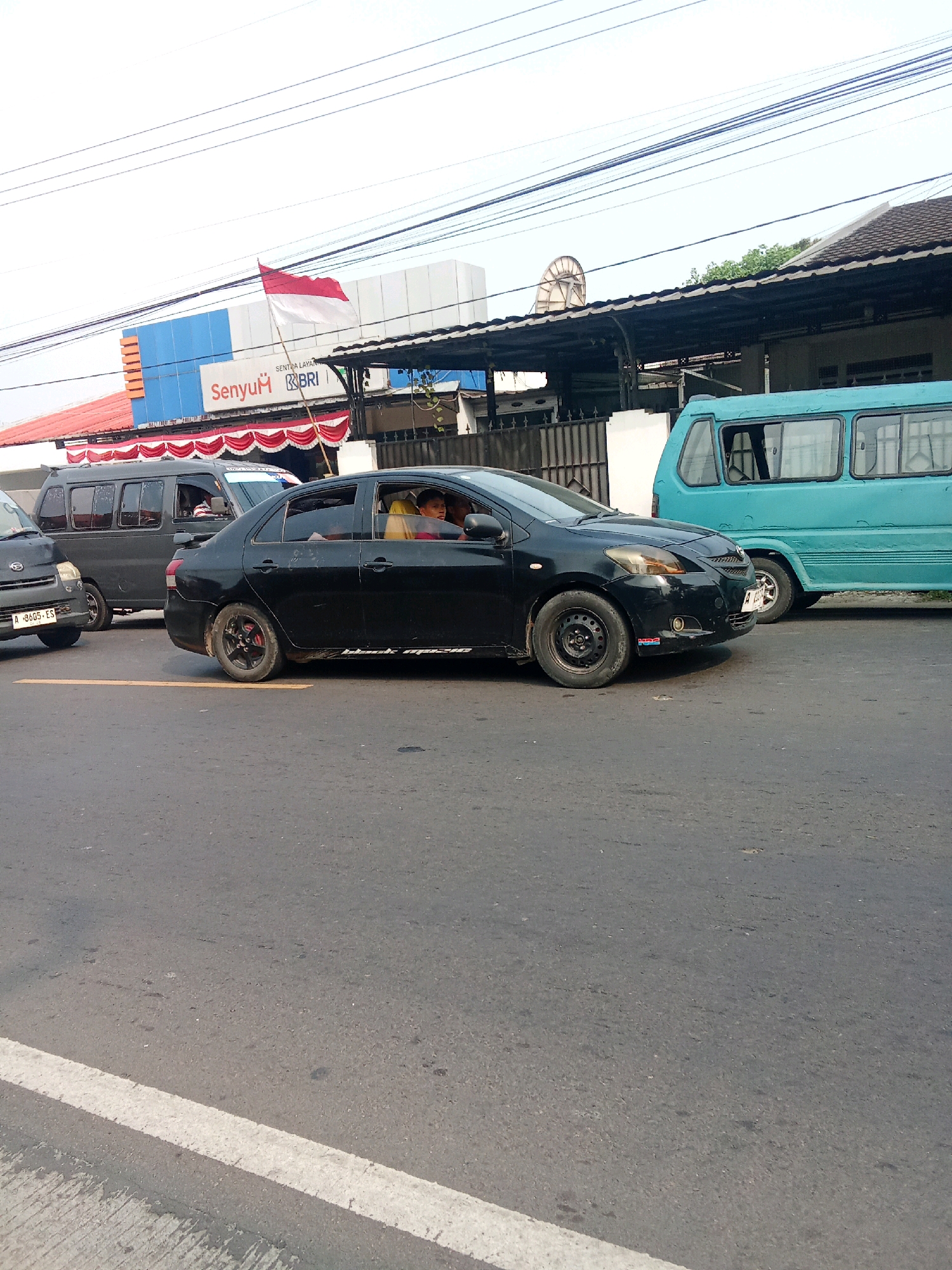 This image depicts a street scene in Warunggunung, Indonesia. In the foreground, a black sedan is stopped in traffic. The driver, a man wearing a yellow shirt, is visible behind the wheel. A passenger in the back seat is also visible. To the left of the black sedan, a dark gray van is also stopped. Further down the street, a turquoise bus is driving away from the viewer. In the background, a building with the text "Senyum BRI" and an Indonesian flag are visible. The sky is overcast. The road is asphalt, with a white line marking the lane.