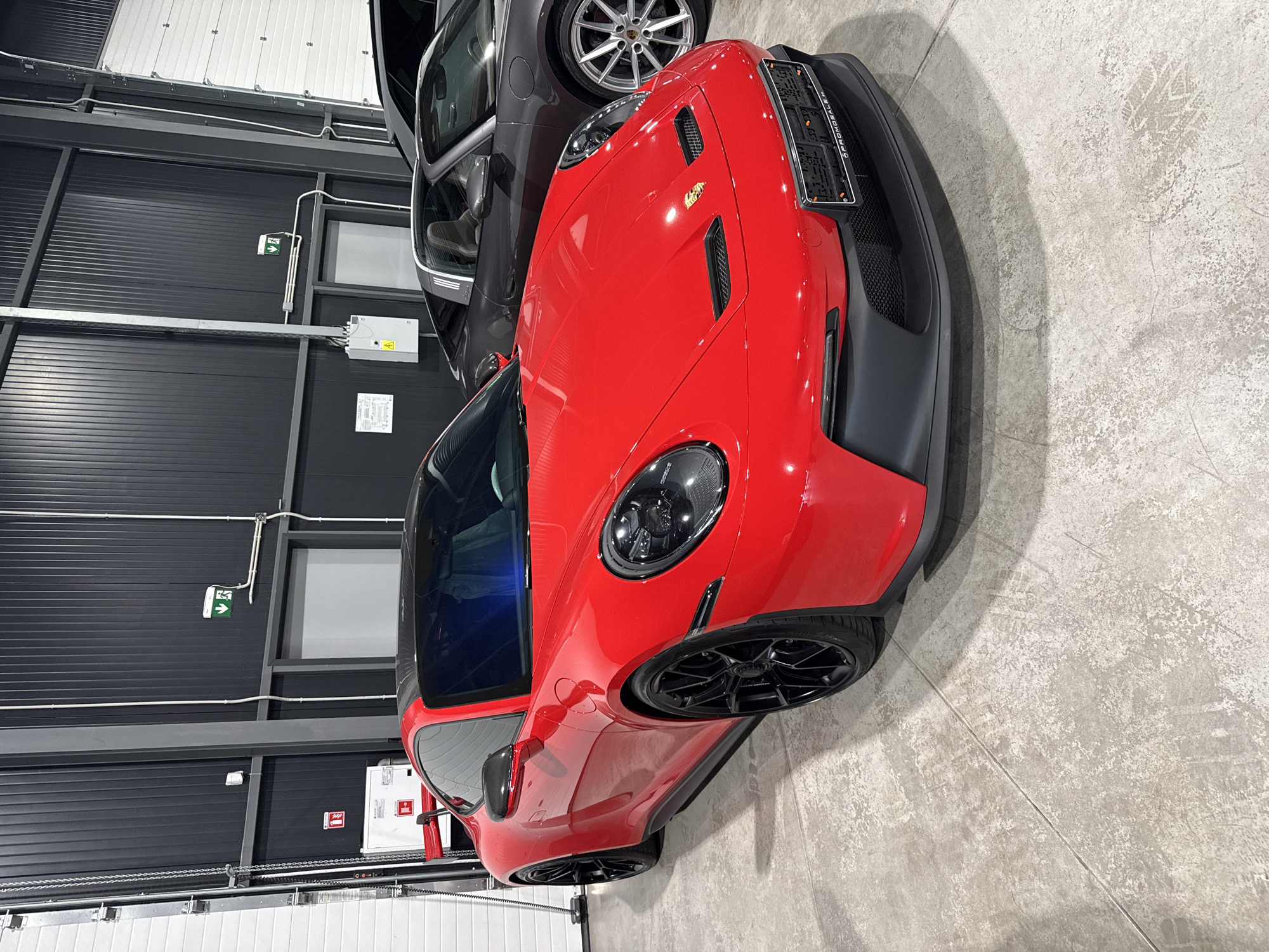 A bright red Porsche 911 GT3 is parked indoors on a grey concrete floor. The car is positioned at an angle, with its front slightly turned towards the viewer. It has black wheels, a black roof, and a prominent rear spoiler. To the right of the red Porsche, a grey Porsche Targa is partially visible. The background consists of a dark corrugated metal wall with white garage doors on the right side and a white rolling door on the left. There are some electrical conduits and signs on the wall, including a green directional sign with arrows. The lighting appears to be artificial and evenly distributed. No people are visible in the image. The scene is likely a car dealership or a private garage in Plovdiv, Bulgaria.