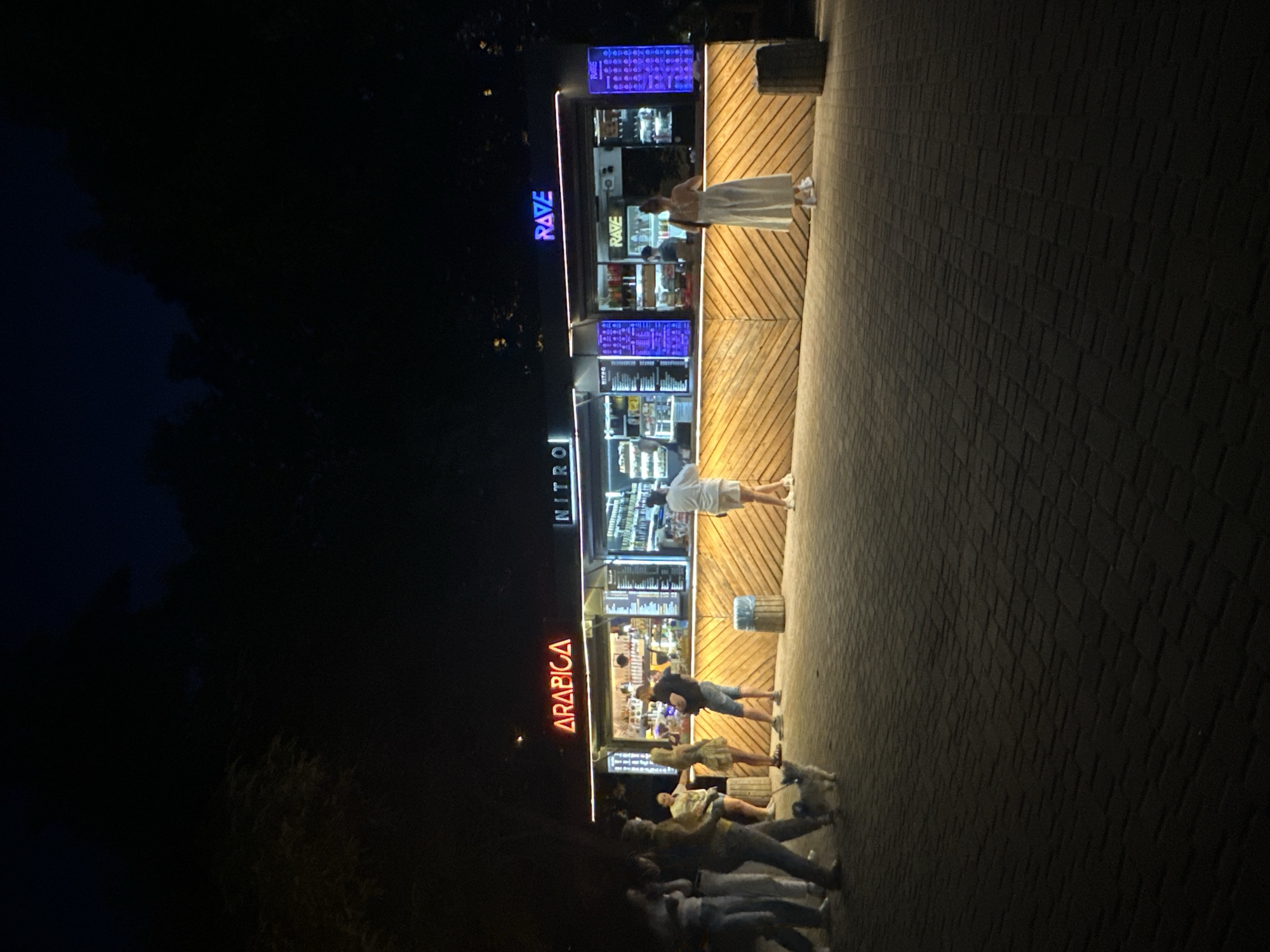 The image depicts an outdoor scene at night in Odesa, Ukraine, likely near the sea or a park, given the trees in the background. A brightly lit, modern food stand or kiosk is the central focus. The kiosk features three distinct sections, each with illuminated signage: "ARABICA" in red neon, "NITRO" in white neon, and "RAVE" in blue neon. The kiosk's facade is constructed of light-colored wooden planks arranged in a chevron pattern, illuminated from above.
Several people are visible. On the left, a group of four individuals, two men and two women, are standing and walking on a paved walkway. One woman appears to be holding a leash attached to a small, light-colored dog. In the center, a man is standing at the "ARABICA" counter, and a woman in a patterned dress is looking at the menu. Further to the right, a person in white attire, likely a woman, is at the "RAVE" counter. Behind the counter of the "RAVE" section, a person is partially visible, seemingly an employee.
The ground is paved with dark interlocking bricks, and two cylindrical trash receptacles are placed in front of the kiosk. The overall atmosphere is lively and urban, suggesting a popular gathering spot in Odesa during the evening.