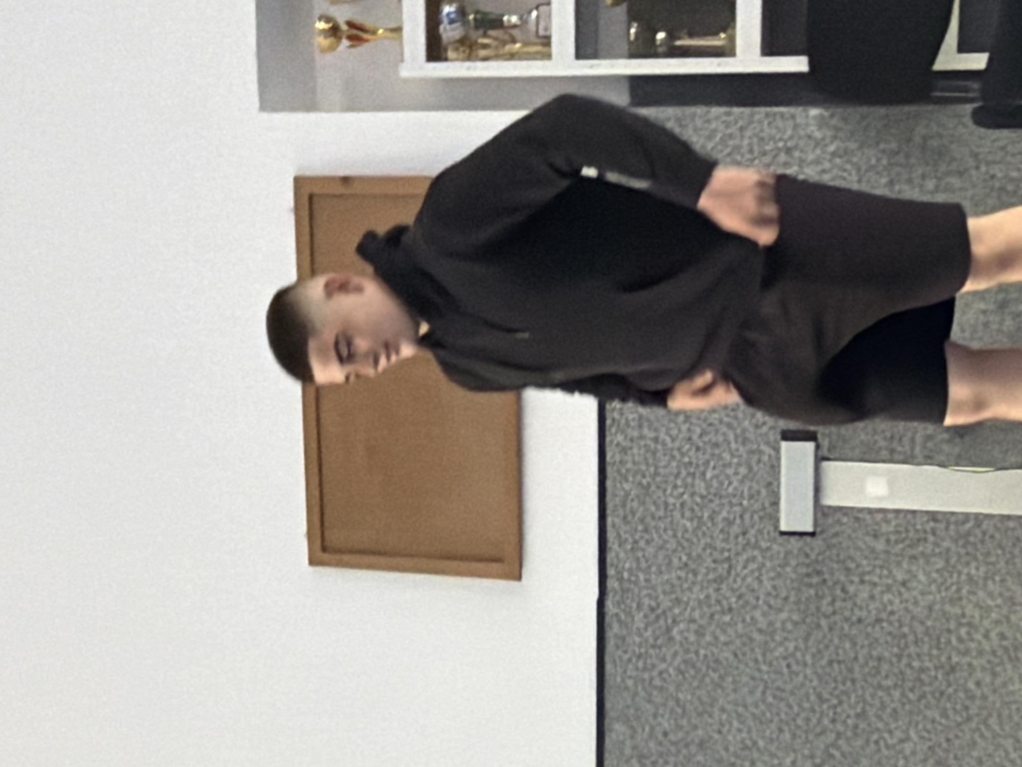 A young man with a buzzcut is standing in what appears to be an indoor space with gray, speckled carpet. He is wearing a black hoodie and dark shorts. Behind him, on the left, is a white wall with a brown, framed object hanging on it. In the upper background, there is a shelf containing what seem to be trophies. The location appears to be in a building, possibly a school or gymnasium, but there are insufficient details to specify if it is in Panagyurishte, Bulgaria.