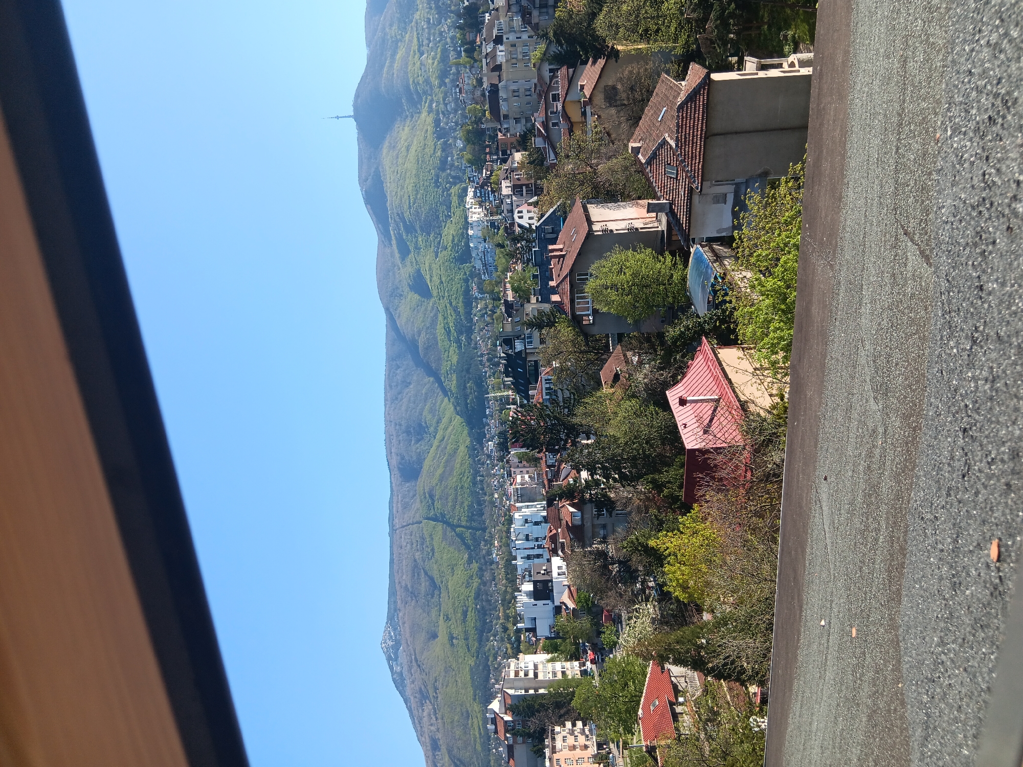 This media file captures a panoramic view of a residential area situated at the base of a verdant mountain range, likely Vitosha mountain overlooking Sofia, Bulgaria. The scene is bathed in bright daylight under a clear, cloudless blue sky, suggesting pleasant weather during spring or early summer.
In the foreground, a coarse-textured, dark gray surface, possibly a flat rooftop or a wide ledge, occupies the bottom right of the frame. A wooden structure, possibly a window frame or part of a building edge, appears on the far right, partially obscuring the view.
The midground showcases a sprawling residential neighborhood. Numerous houses and apartment buildings of varying architectural styles are visible, many featuring characteristic red or terracotta tiled roofs. The area is rich with lush green trees and foliage, interspersed among the structures, indicating a well-treed urban environment. One building in the midground has a distinctive blue-covered structure next to it, potentially a swimming pool or greenhouse.
Dominating the background is a large mountain, densely covered with vibrant green deciduous forest, highlighting the season. A tall, slender communication or television tower stands prominently on a distant ridge of the mountain, slightly to the left of the center, a known landmark for the area.
No human activity or people are visible. The image conveys a serene and natural integration of urban life with the surrounding mountain landscape. There is no visible text within the image.