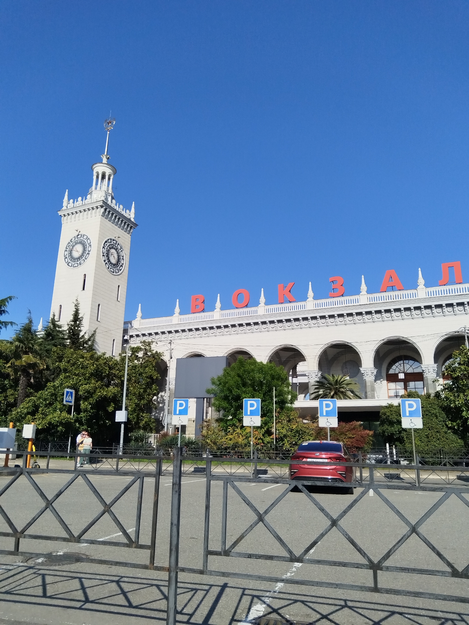 The image shows the Sochi Railway Station in Russia. The building is white with large red Cyrillic letters spelling "ВОКЗАЛ" on the front, which translates to "Station." A tall clock tower stands on the left side of the building. The station has arched openings on its facade, and there are parking signs indicating handicapped parking in front. A red car is parked in one of the spaces. A metal fence separates the parking area from the viewer. Trees are visible around the station. A person is seen standing near a pedestrian crossing sign on the left. The sky is clear and blue.