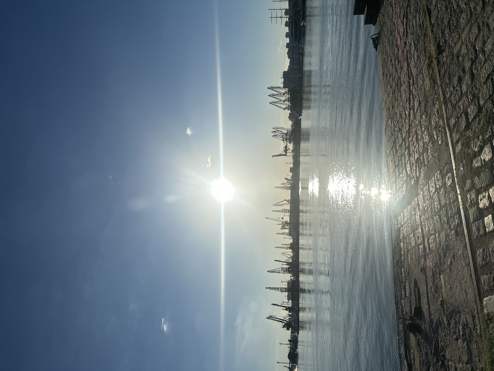 The image depicts a harbor scene at sunset or sunrise. A large body of water occupies the foreground and middle ground, reflecting the bright sun. The water appears calm with slight ripples. In the background, a silhouetted cityscape is visible, dominated by numerous industrial cranes lining the waterfront. Buildings are discernible behind the cranes. A tall ship with sails is moored on the far right side of the harbor. The sky is a clear blue with a few wispy clouds and a prominent lens flare from the sun. The immediate foreground is a cobblestone pier with some dark objects and metal fixtures. The overall impression is one of industrial activity within a port environment bathed in the warm light of the sun.
