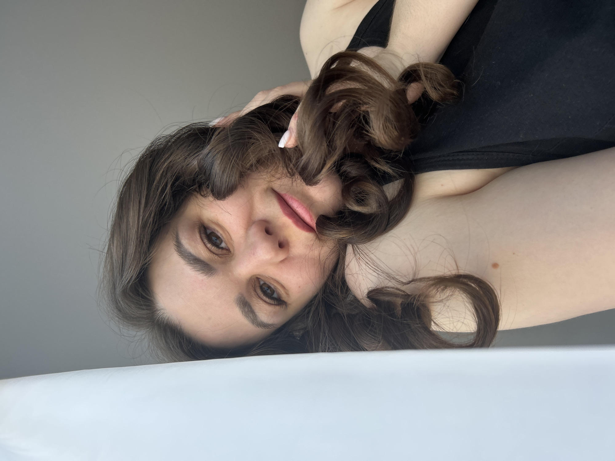 A woman with brown curly hair and fair skin is lying down, facing the camera. She is wearing a black tank top. Her left hand is near her head, partially obscuring her face. The background is a plain light gray wall. The foreground is white. There is no indication of the city of Chaikovsky, Russia, or any other specific location.