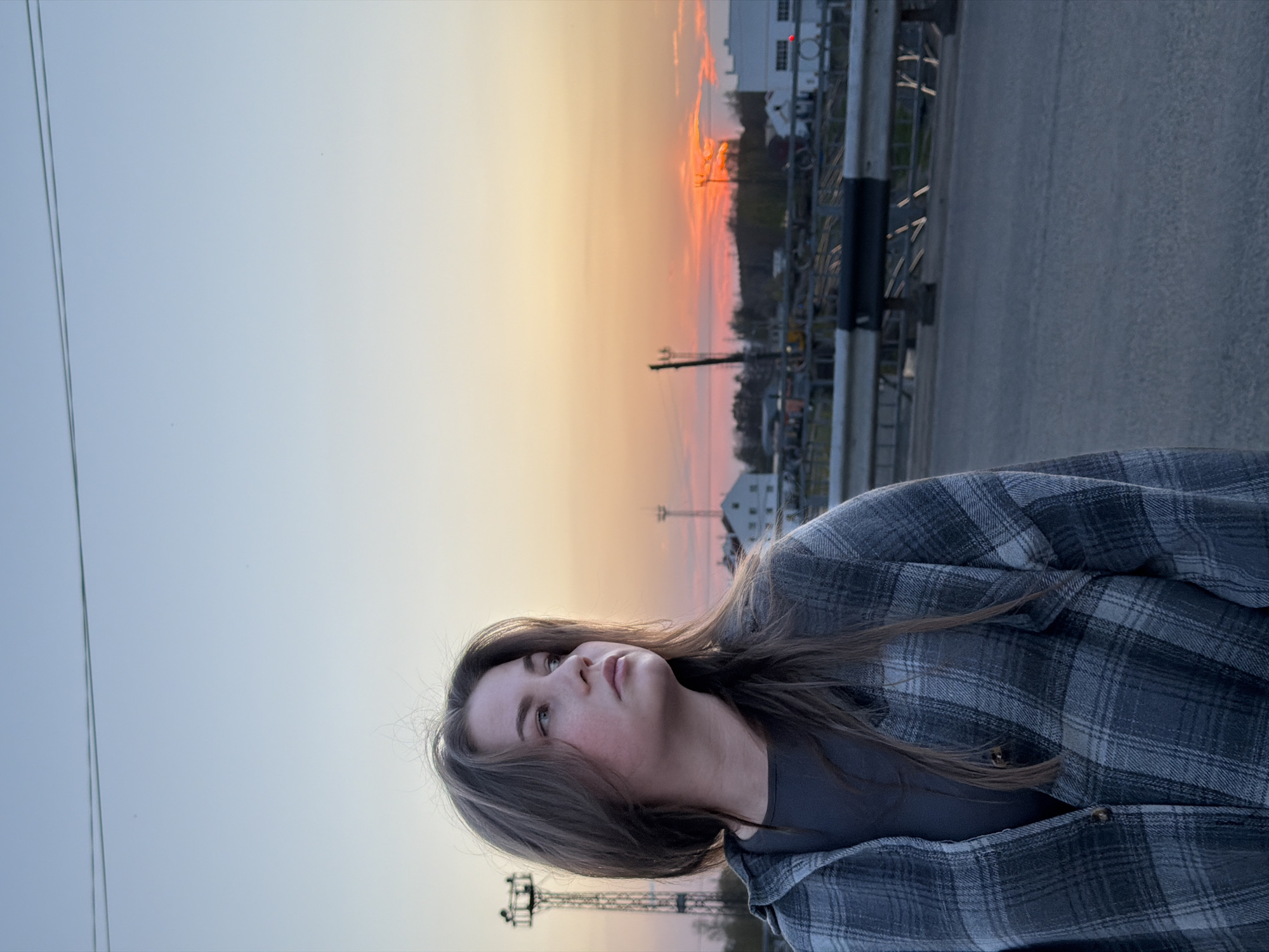 A woman in a gray flannel shirt stands on a paved road, facing the sunset in Chaikovsky, Russia. The sky transitions from light blue to orange, with a building visible in the distance. Metallic structures and railway tracks are present. The woman looks up, her face illuminated by the setting sun.