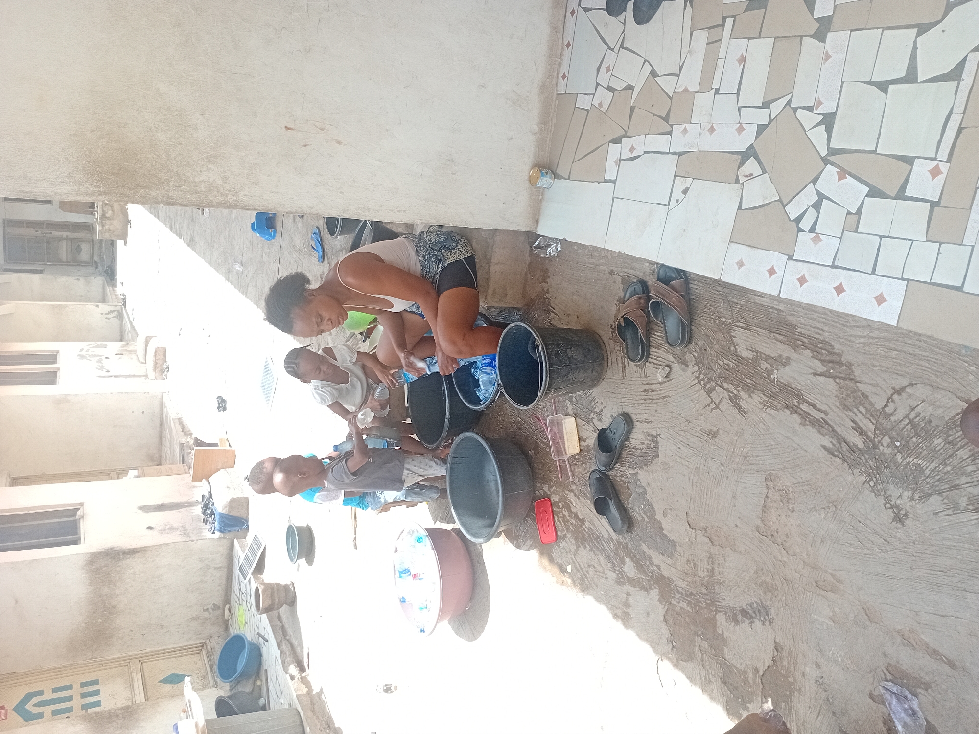 An outdoor scene captures a group of people, primarily a woman and several children, engaged in a washing activity. The image is oriented vertically but depicts an outdoor courtyard or alleyway bathed in bright daylight.
In the foreground, a young woman with dark hair, wearing a light tank top and patterned shorts, bends over a large black plastic tub, appearing to wash items. She is focused on her task. Several children are around her; one small child is visible close to her, and another young boy, bald-headed, stands near a brown tub filled with plastic water bottles, looking at the washing activity. Other children are partially visible in the background.
Numerous objects related to washing are scattered on the concrete and tiled ground. These include several large plastic tubs and buckets of various colors (black, brown, blue), some filled with water and plastic bottles. A pink washing brush, a yellow rectangular item (possibly soap), and a red plastic container are placed on the ground near the tubs. Multiple pairs of slippers and sandals in brown, black, and blue are also visible.
The ground consists of a rough concrete surface and, in the top right section, a decorative area paved with irregularly shaped beige and white tiles, some featuring small red diamond patterns. In the background, light-colored buildings with windows and doors line the space, suggesting a residential environment. One door has a distinctive blue and white emblem. The strong sunlight casts clear shadows, indicating a bright, sunny day. The overall atmosphere suggests a communal or household chore being performed outdoors.
Visible text includes partially legible "AQUA" on a blue plastic water bottle.