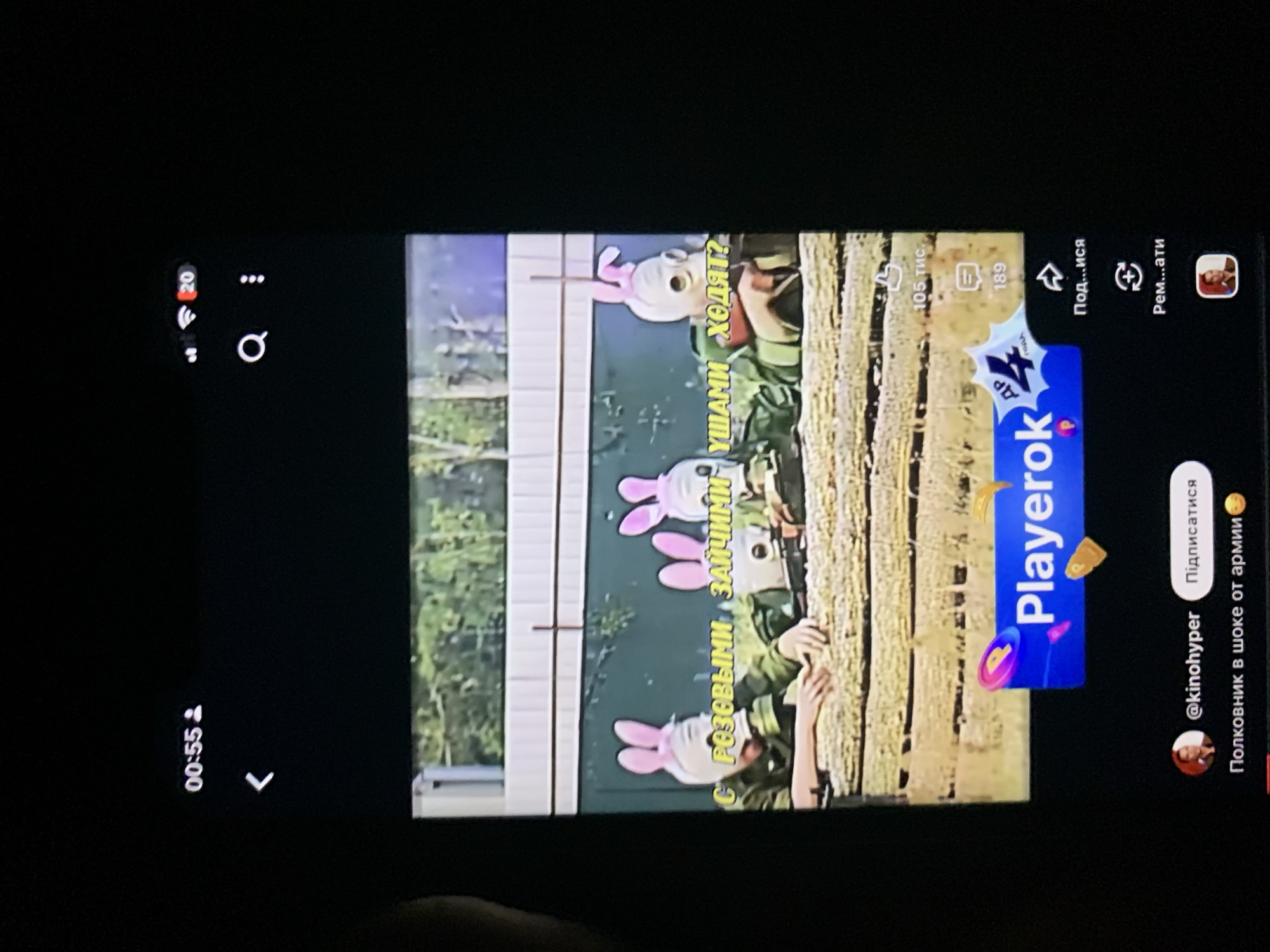 The image shows a close-up of a mobile phone screen displaying a video with a humorous theme. The video features three individuals wearing gas masks and pink bunny ears, dressed in military-style uniforms. Yellow text overlays the video, which appears to be in Cyrillic script.
The top left corner of the phone screen displays "00:55," indicating the video's current playback time. To the right of this is a small user icon. A white arrow pointing left is also visible below the timestamp.
The video content itself shows the three individuals, likely soldiers, in an outdoor setting with trees and a fence in the background. They are positioned behind a hay bale or similar material. The yellow text on the screen translates to "With pink bunnies, they walk?"
Below the video, a blue banner with the word "Playerok" is displayed, accompanied by animated icons. Further down, a profile picture of a person is visible next to the username "@kinohyper" and a "Subscribe" button. Below this, more Cyrillic text reads "Colonel in shock from the army" with a smiling emoji.
On the right side of the screen, several icons are present: a thumbs-up icon with "105 тис." (105 thousand) below it, a comment icon with "189" below it, a share icon, and a refresh icon.
The provided image does not contain any visual information that would allow for the determination of its location in Zhmerynka, Ukraine, or any other specific geographical location.