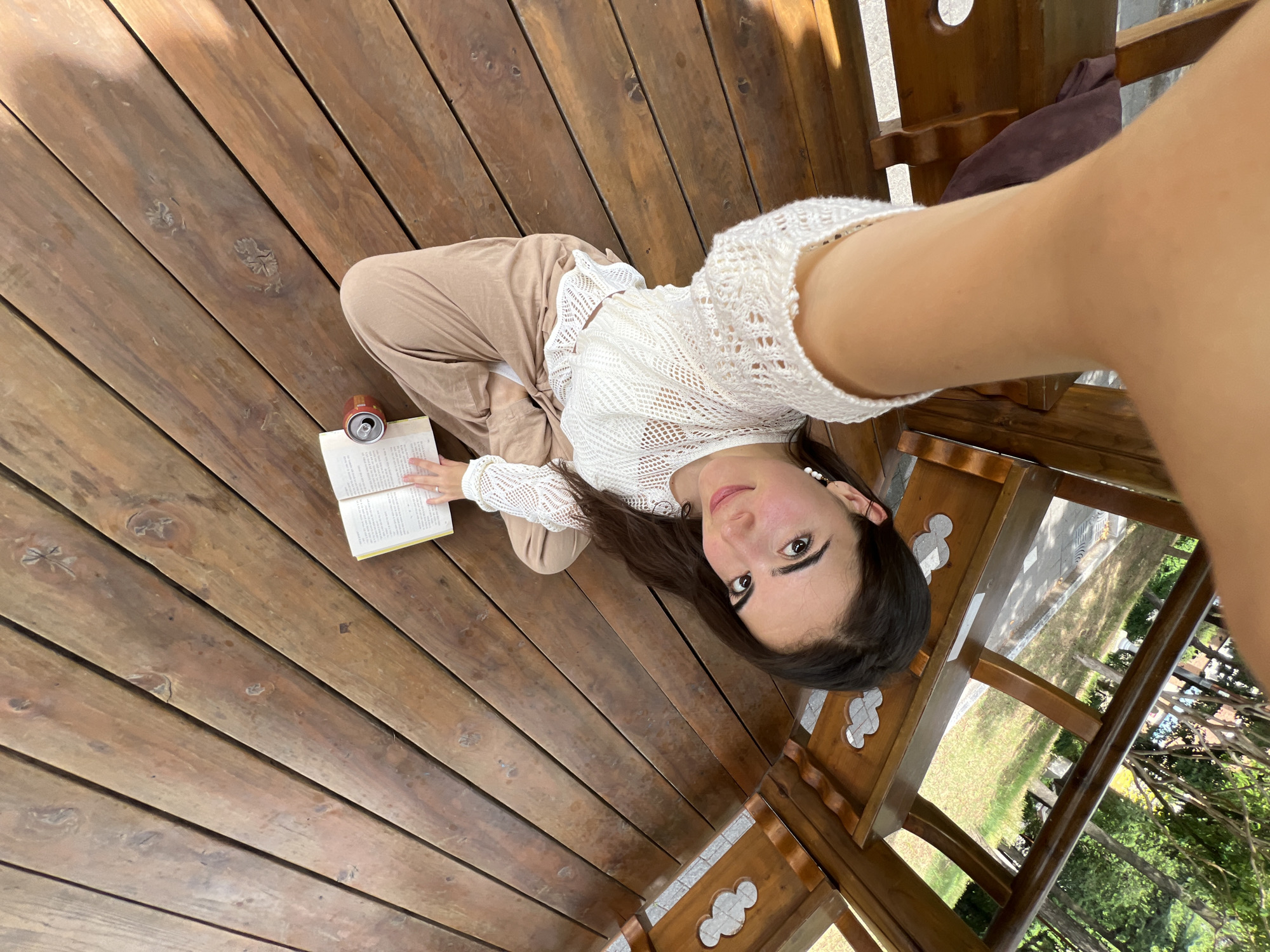 A young woman is sitting cross-legged on a wooden deck, holding an open book. She is wearing a white, crocheted top and light brown pants. A soda can is placed next to the book. The wooden deck appears to be part of an outdoor structure, possibly a gazebo or covered patio, with decorative wooden elements visible. Beyond the structure, a grassy area and trees can be seen, suggesting an outdoor setting. The image is taken from a high-angle perspective, looking down at the woman. The woman's face is turned towards the camera, and she is looking directly at it. Her expression is neutral. The lighting suggests it is daytime, with sunlight casting shadows on the wooden deck. This scene is set in the city of Daegu, South Korea.