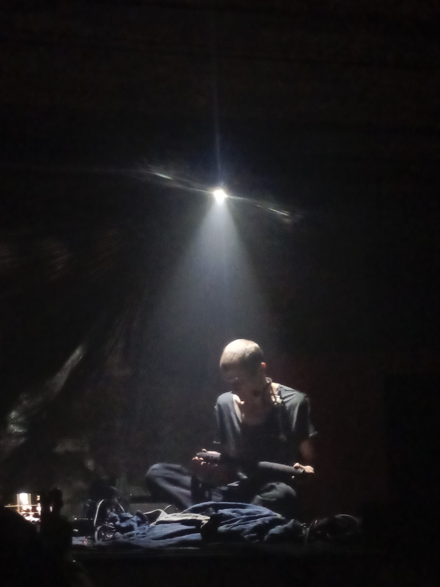 A person is sitting cross-legged in a dark environment, illuminated by a single beam of light from above. The person is wearing a dark t-shirt and has a short haircut. They appear to be holding or interacting with an object in their lap. In the foreground, there are some folded dark fabrics. The overall environment is dark with some fuzzy textures visible in the background. No specific details about the location in Moscow, Russia, are discernible from the image.