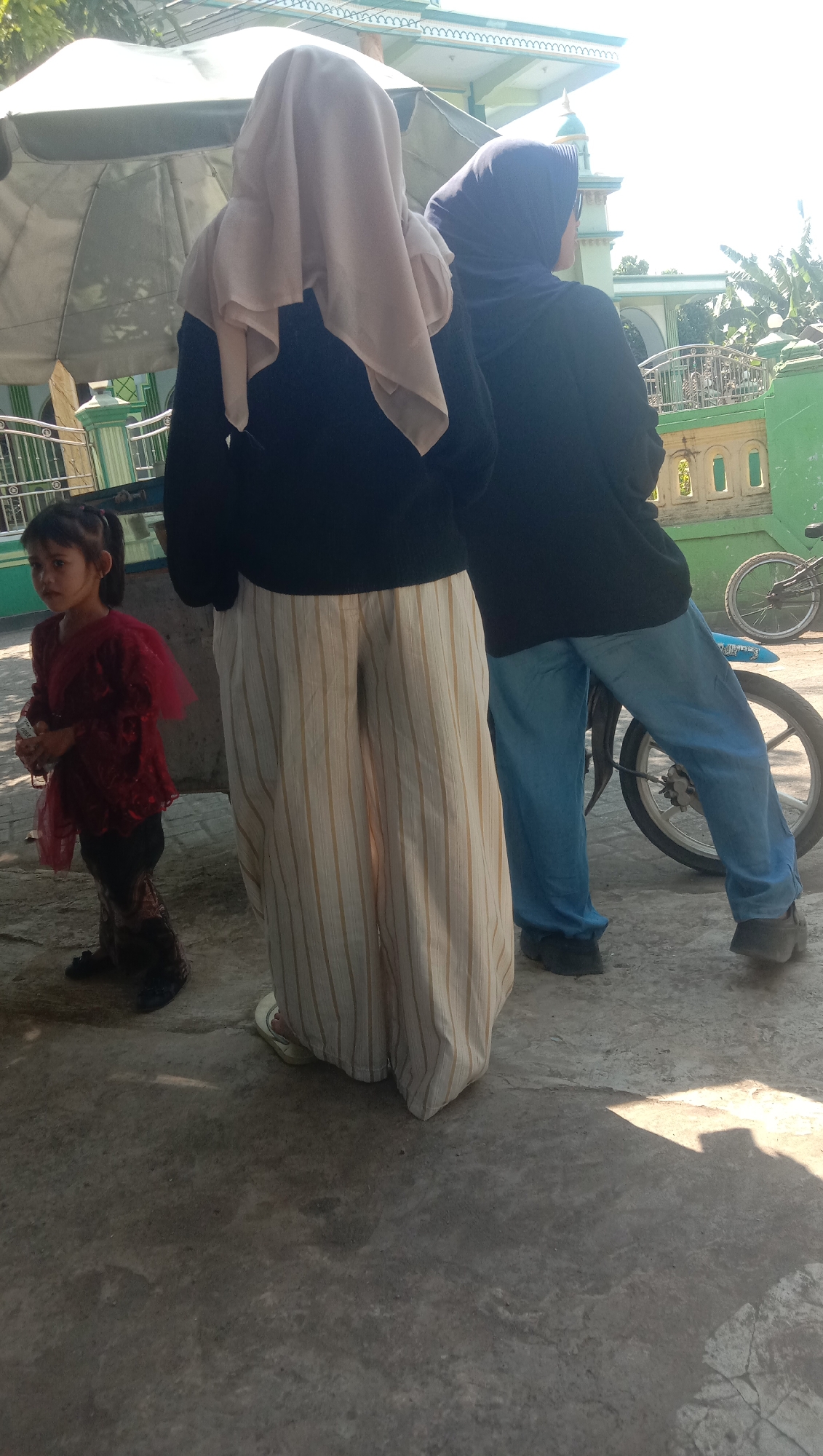 In Ngempit, Indonesia, on a paved surface, three individuals are standing outdoors. The subject on the left is a young girl with dark hair pulled into a ponytail, wearing a maroon top with ruffled sleeves and dark pants. She is holding a small white object. To her right, a person wearing a light-colored hijab and a dark top is partially visible. Their lower half is obscured by a person in the center, who is wearing a dark hijab and a dark top, with light blue jeans. This person is leaning against a motorcycle with a white rim and spokes. In the background, a building with green accents and a tiled roof is visible, along with foliage and a bright sky. A large umbrella is positioned above the two individuals wearing hijabs. The ground is concrete with some cracks and shadows cast by the sun.