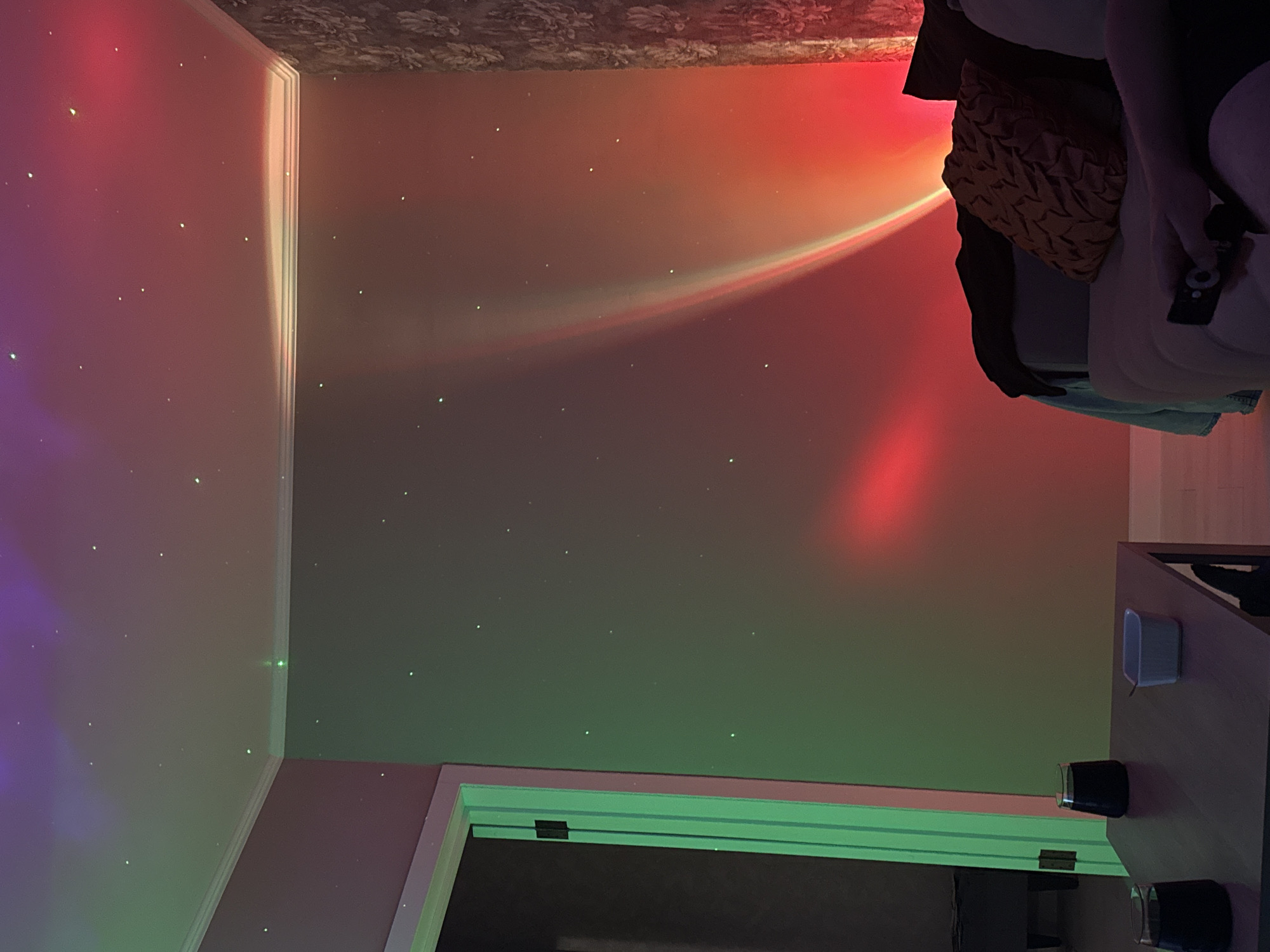 The image depicts an indoor residential living space, likely a living room or den, with its exact city and location indeterminable. The room is illuminated by a dynamic light projector, casting a nebula-like array of purple and green hues across the ceiling and upper left wall, accompanied by numerous small, green 'star' dots distributed across all visible surfaces. A prominent, arcing beam of light, transitioning from red to orange and yellow, originates from the lower right and stretches diagonally up the right wall.
A light-colored sofa occupies the right side of the frame, draped with a dark blanket and featuring a textured, orange-brown throw pillow. A person is partially visible on the sofa, with their right arm and hand holding a black remote control, suggesting interaction with entertainment or lighting systems. In the foreground, a dark, rectangular table holds two dark, translucent glasses and a small white rectangular dish containing a spoon or small utensil.
The left wall is plain and transitions into a light green-framed doorway leading to a darker room. The rightmost wall features a patterned floral wallpaper. White crown molding defines the junction of the walls and ceiling throughout the visible area.
