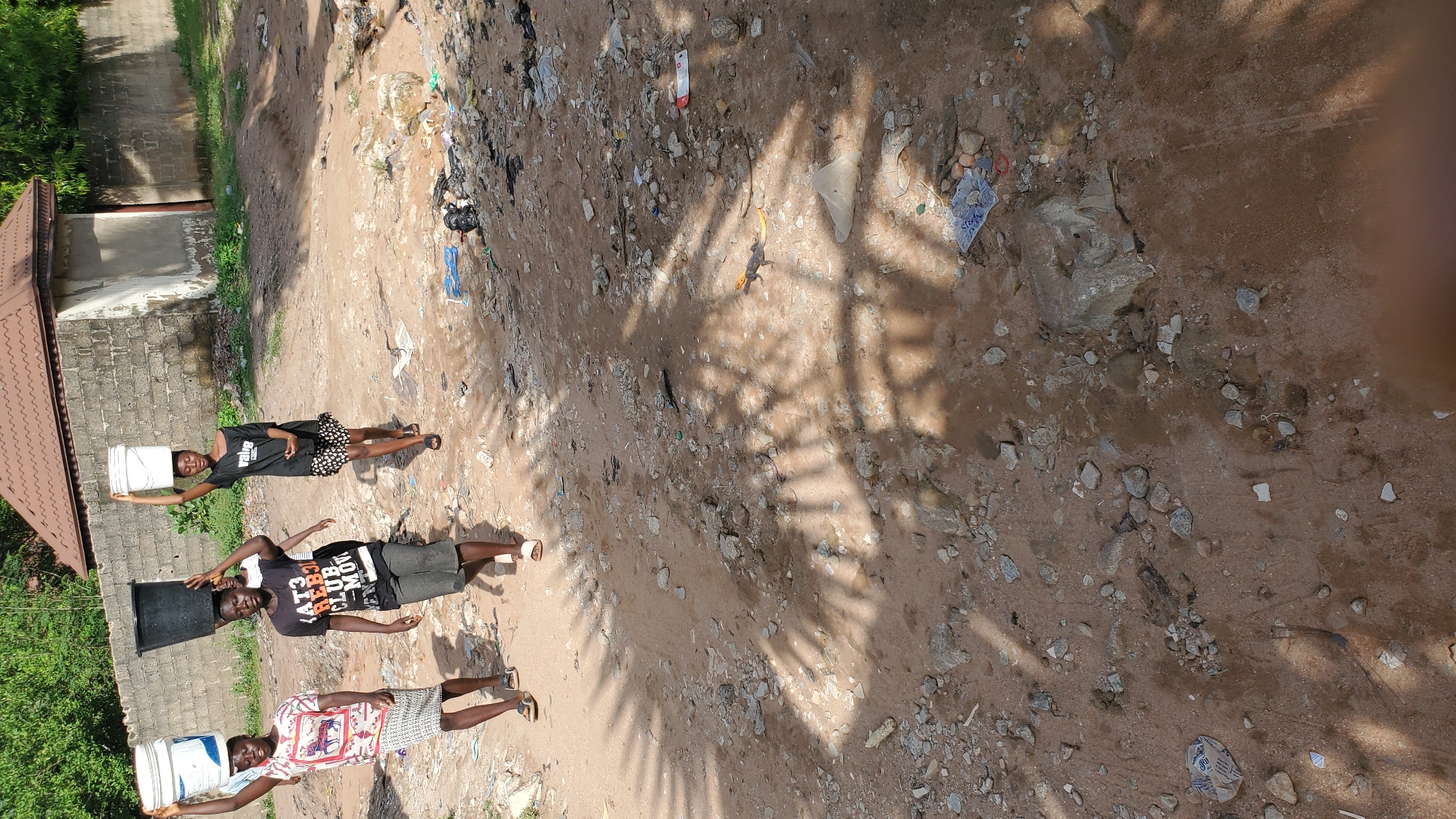 In Jalingo, Nigeria, four young people walk on a dirt road littered with debris. The three in the foreground are carrying buckets of water on their heads. The person on the far left wears a patterned shirt and a patterned skirt. Next to them, a person in a black t-shirt with a graphic design and grey shorts walks with a black bucket on their head. The third person, to the right of the second, wears a black t-shirt and a patterned skirt, also carrying a white bucket. A fourth person, partially visible behind the first, is also carrying a white bucket. In the background, a concrete wall borders the property, with a building structure featuring a brown tiled roof. The ground is sandy with numerous rocks and scattered garbage.