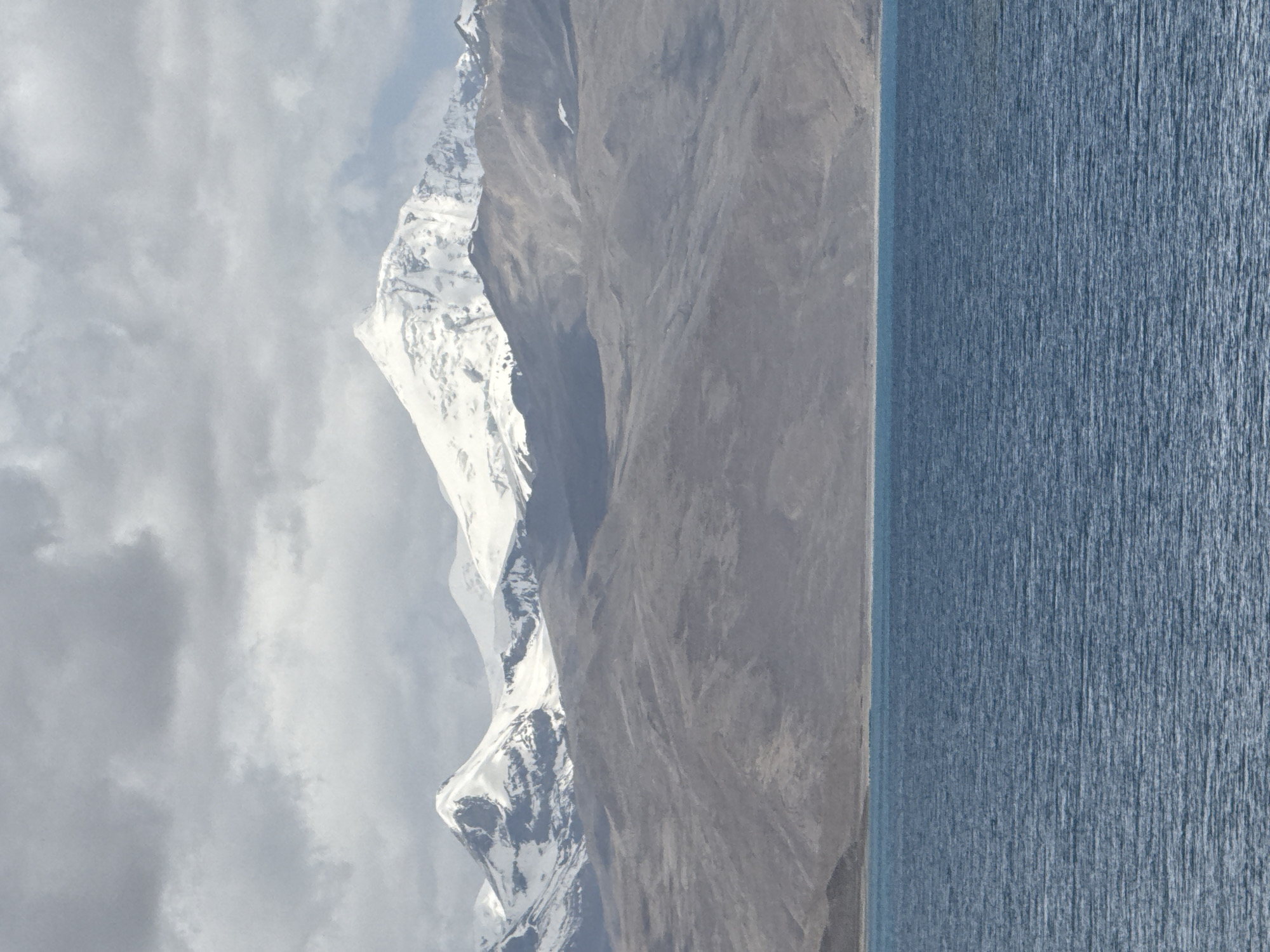 The image depicts a mountain range with snow-capped peaks bordering a body of water. The mountains are primarily grey rock with patches of white snow, and the sky is overcast with grey clouds. The water appears calm with a textured surface. The image appears to be taken at a landscape level. There is no information available to suggest the image was taken in the city of Baidê, China.