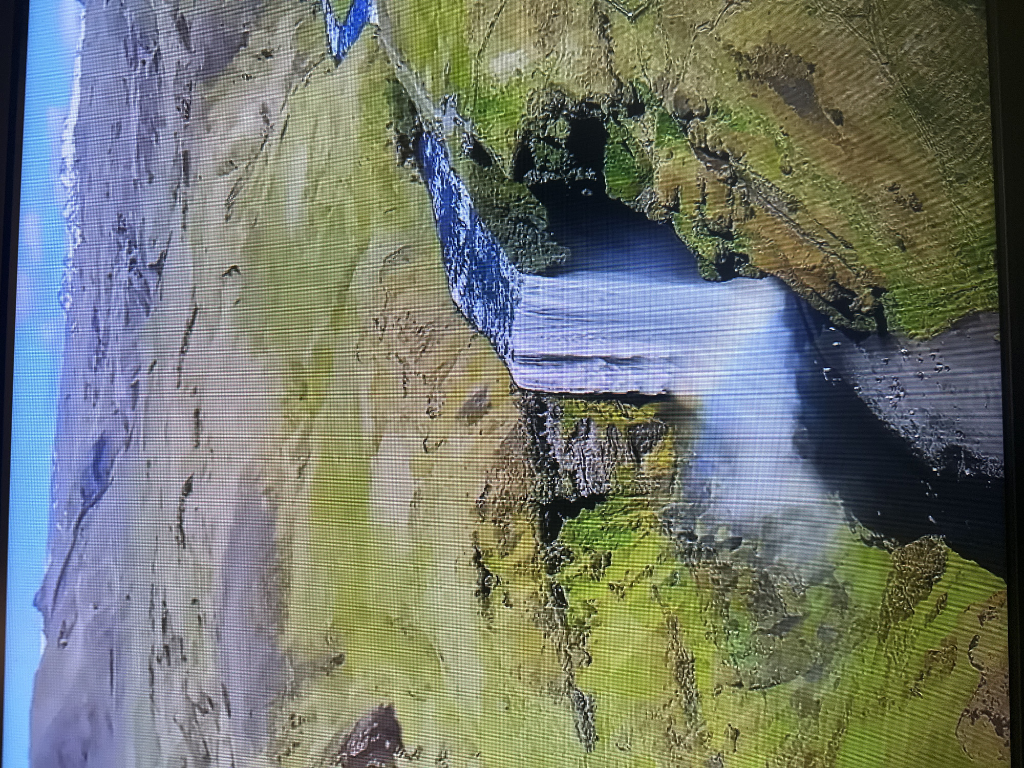 The image is an aerial view of a wide waterfall cascading over a cliff face. The surrounding landscape is characterized by rolling green hills, a winding river, and distant mountains with snow-capped peaks. There is a misty spray at the base of the waterfall, and the water is a deep blue. The scene appears to be natural and undeveloped. The image does not depict any elements that would place it in the city of Kyiv, Ukraine.