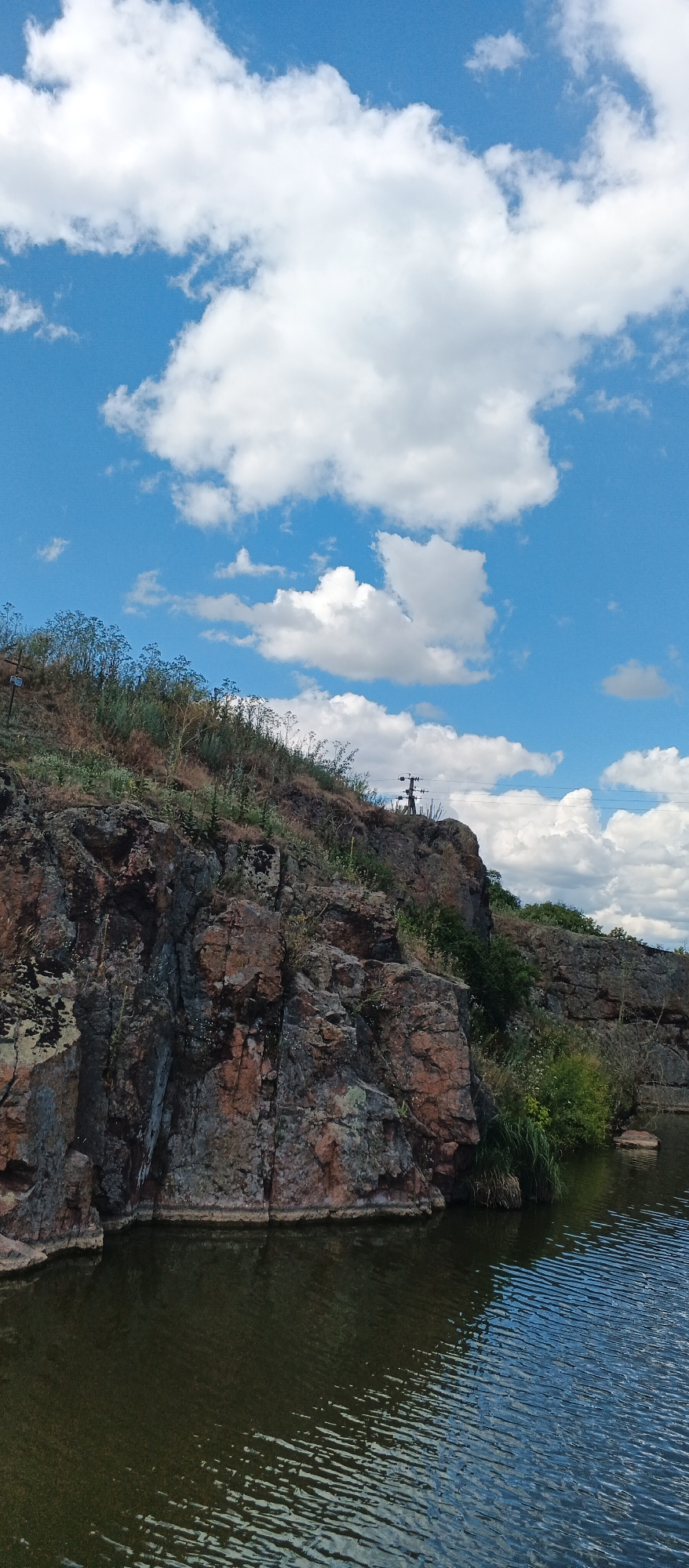 The image depicts a rugged, rocky cliff face overlooking a body of water. The cliff is covered in dry grass and sparse vegetation, with several rocky outcrops and crevices. At the top of the cliff, a person is visible standing with a bicycle. The sky above is a vibrant blue, dotted with fluffy white clouds. The water in the foreground is dark and reflects the sky and the surrounding landscape. The scene appears to be located at the Ternivka quarry in Ternivka, Ukraine, a place known for its natural beauty and recreational opportunities.
