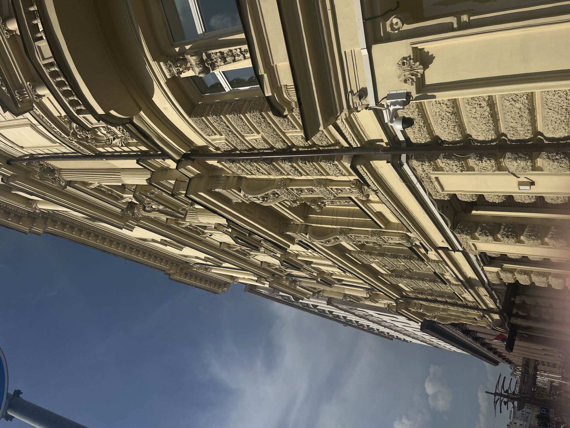 The image shows a detailed view of an ornate building facade in Moscow, Russia, as viewed from below, with the sky in the background. The architecture features classical details such as moldings, decorative cornices, and windows. There is also a security camera present on the building. The sky is blue with some clouds. Street signs, trees, and part of a building are visible at the bottom right, suggesting an urban location.