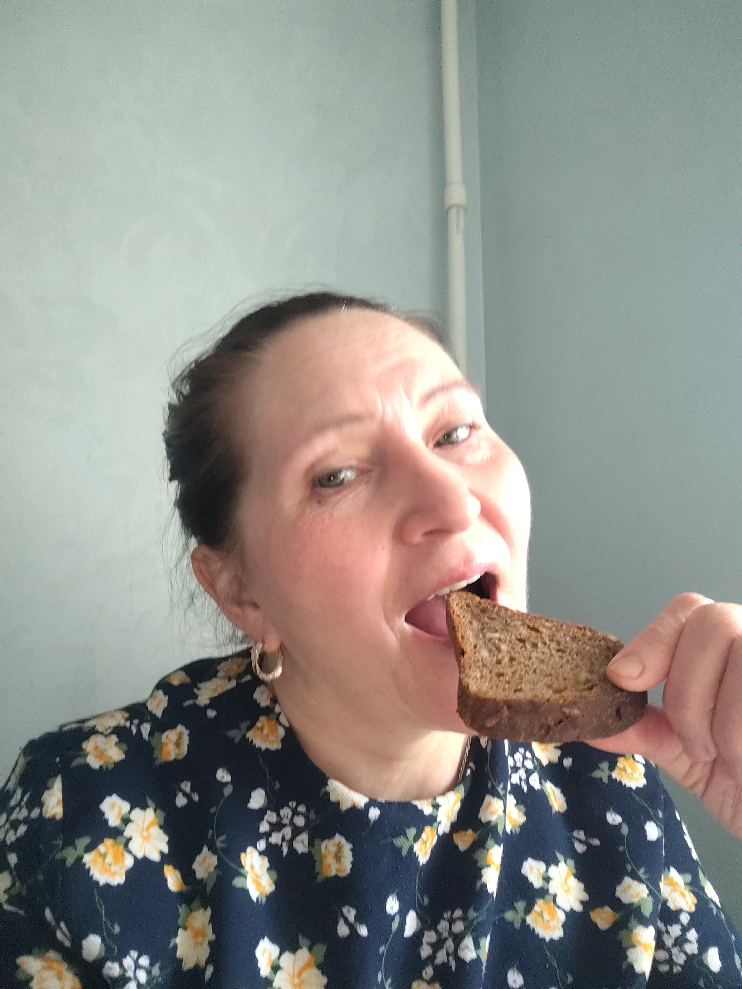 A woman is eating a piece of brown bread. She is wearing a dark blue shirt with yellow and white floral patterns and gold hoop earrings. The background is a light blue-gray wall with a white pipe running vertically along the upper part of the wall. The location is Sarapul, Russia.