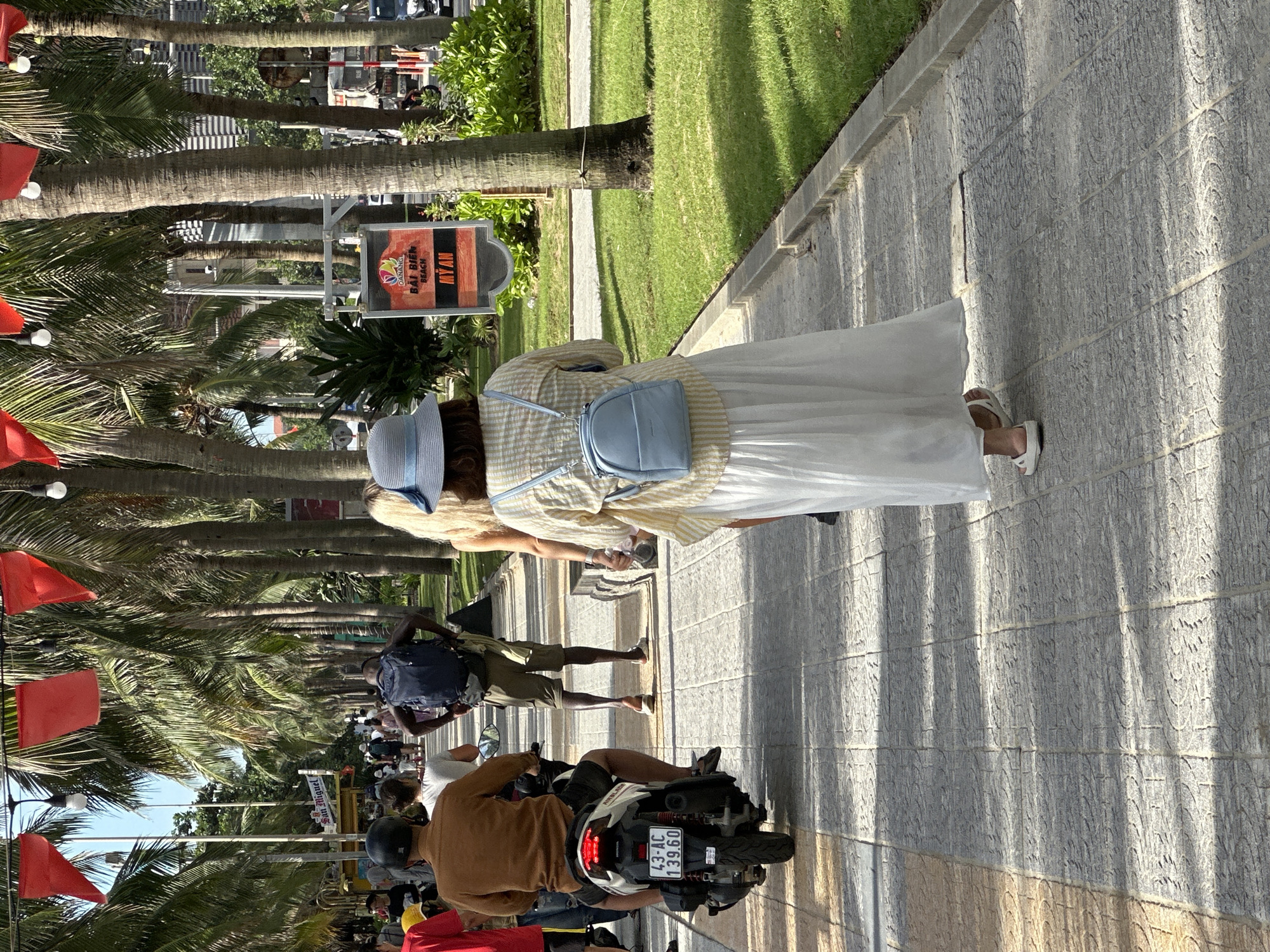 This is a street-level, medium shot of a pedestrian walkway lined with palm trees in Da Nang, Vietnam, specifically along My An Beach. The walkway is paved with light-colored, patterned tiles. On the left, a person on a scooter, wearing a brown shirt and black helmet, is moving away from the camera. The scooter has a license plate reading "43-AC 139.60". Beyond the scooter, several other people are walking.
In the center of the frame, a woman wearing a light blue hat, a yellow and white checkered shirt, and a long white skirt walks away from the camera, carrying a light blue backpack. To her left, another woman with blonde hair is visible from the side, also walking away.
In the background, more palm trees create a dense canopy, with red flags strung between them. A signpost for "DANANG BAI BIEN BEACH MY AN" is visible on the right side of the walkway. The environment appears sunny with shadows cast by the trees. Green grass borders the walkway.